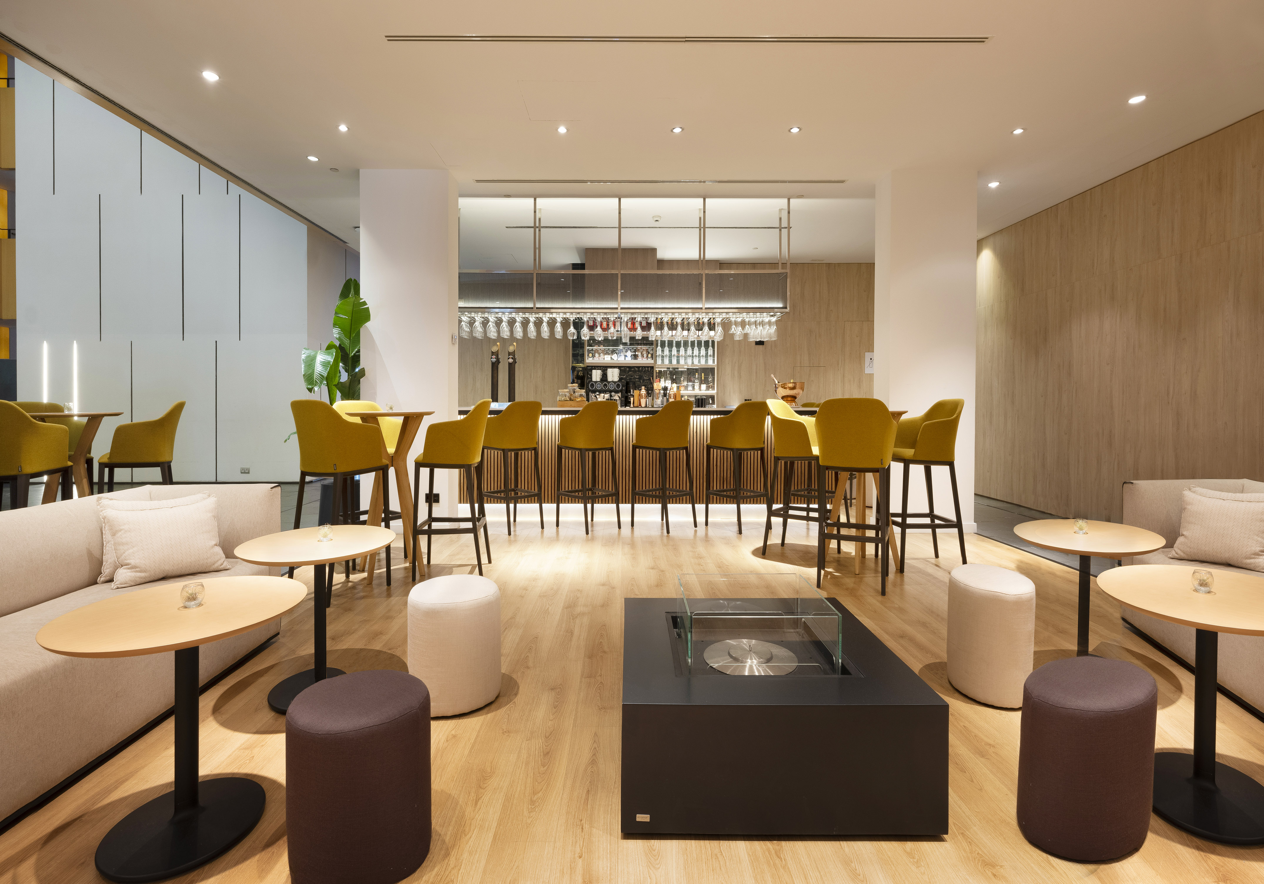 Modern bar lounge area with yellow stools, sofa seating, and a central fire pit.