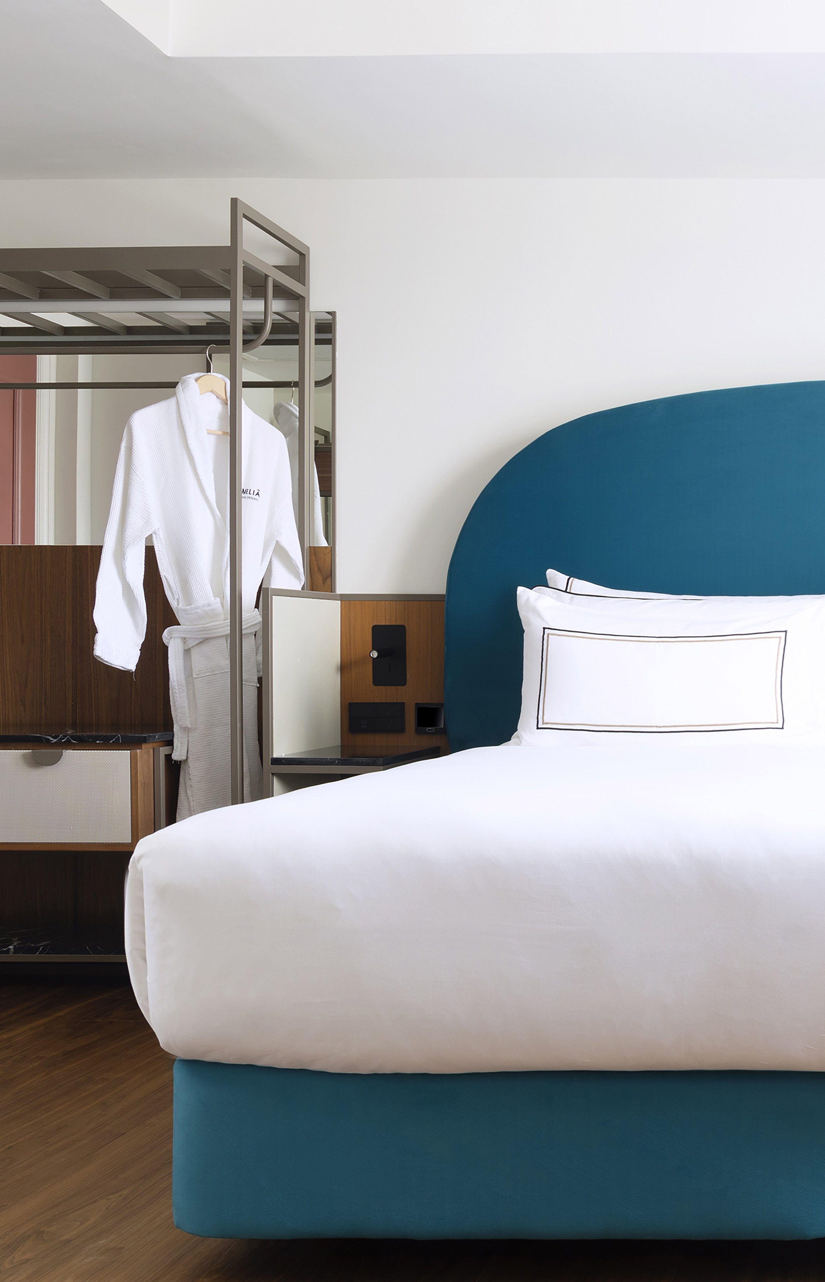 Modern hotel room: teal headboard, white bed, and a bathrobe on a rack.