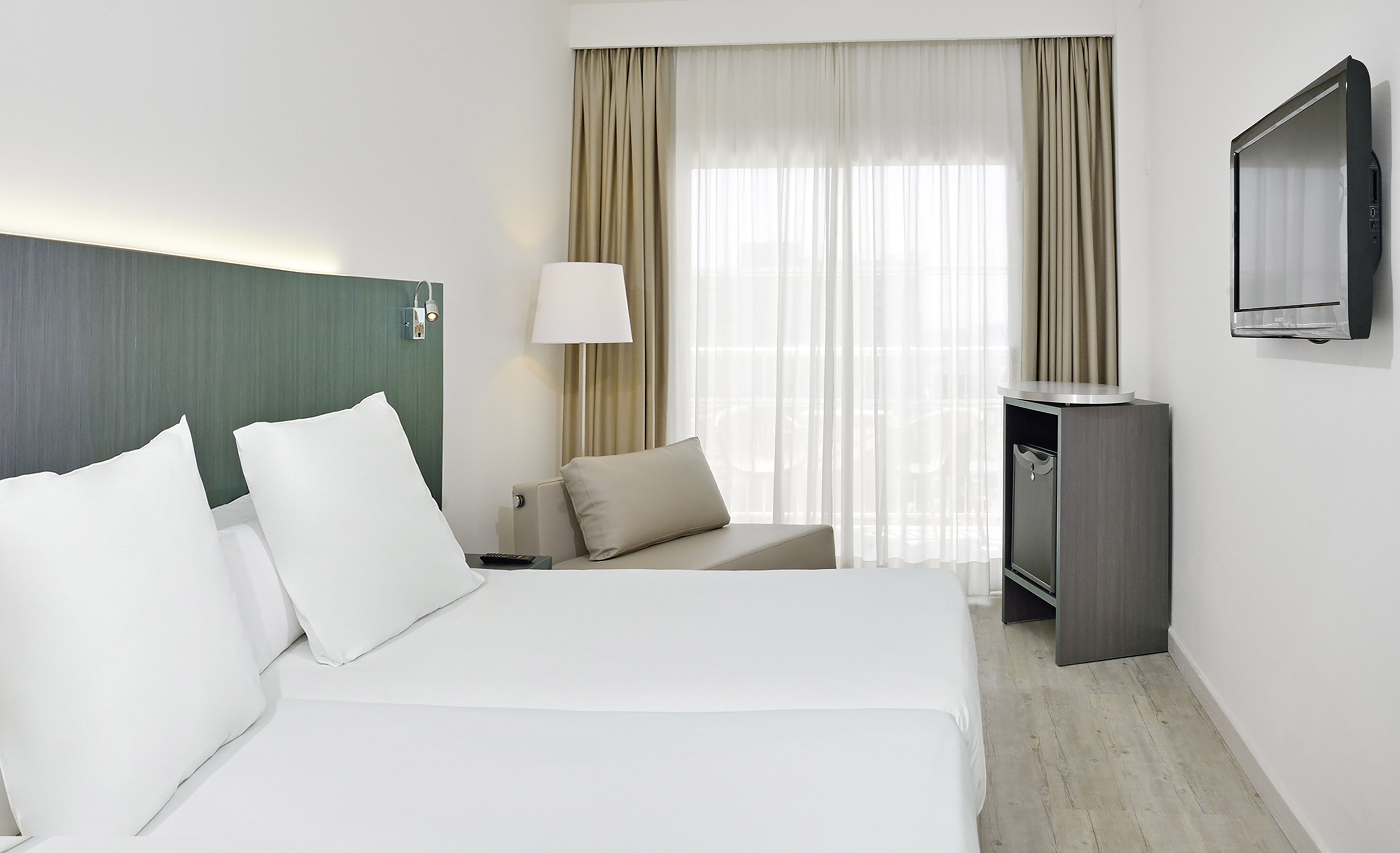 Modern hotel room with twin beds, lounge chair, wall TV, and bright window.