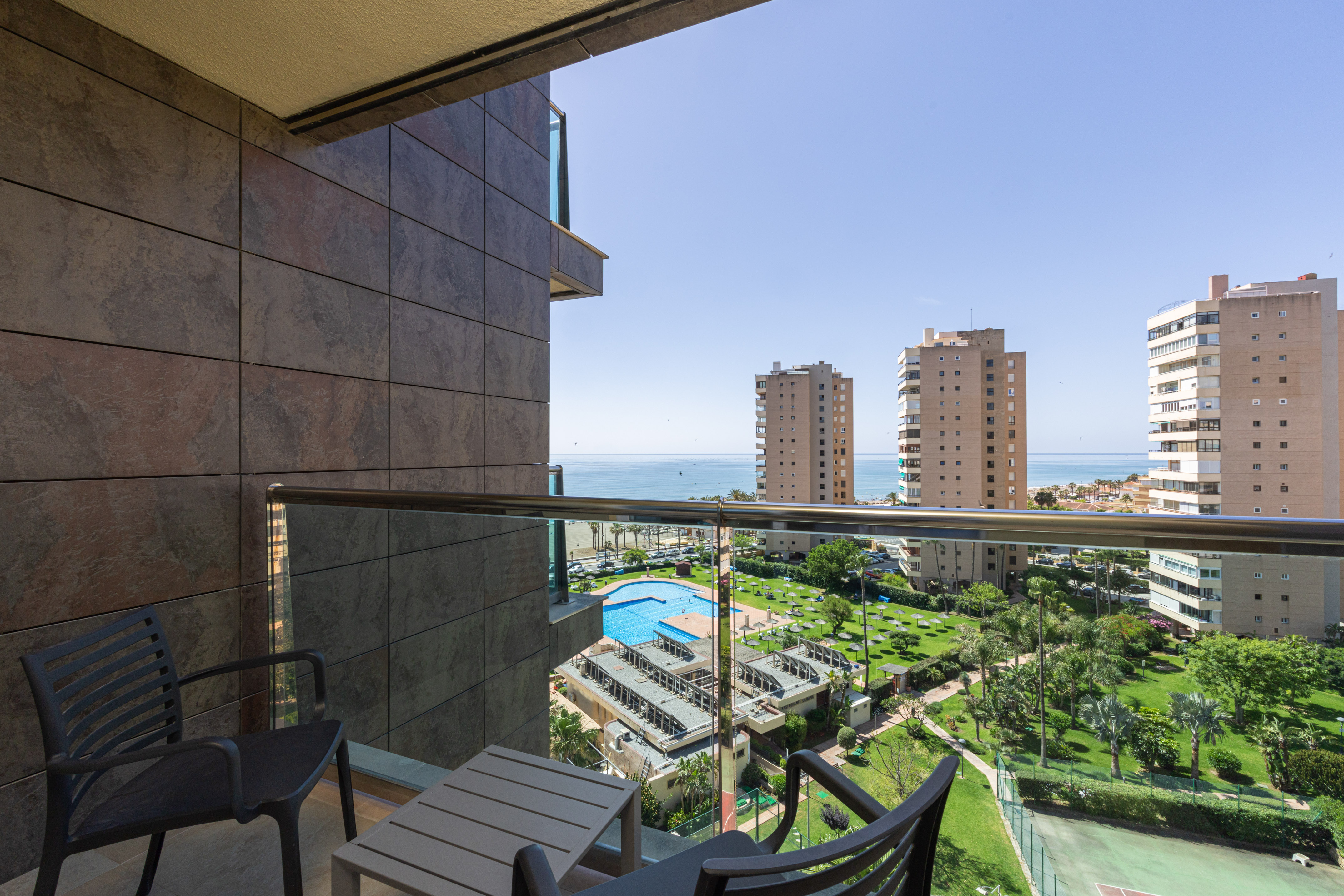 Balcony with clear ocean view, swimming pool, green resort gardens, and buildings.