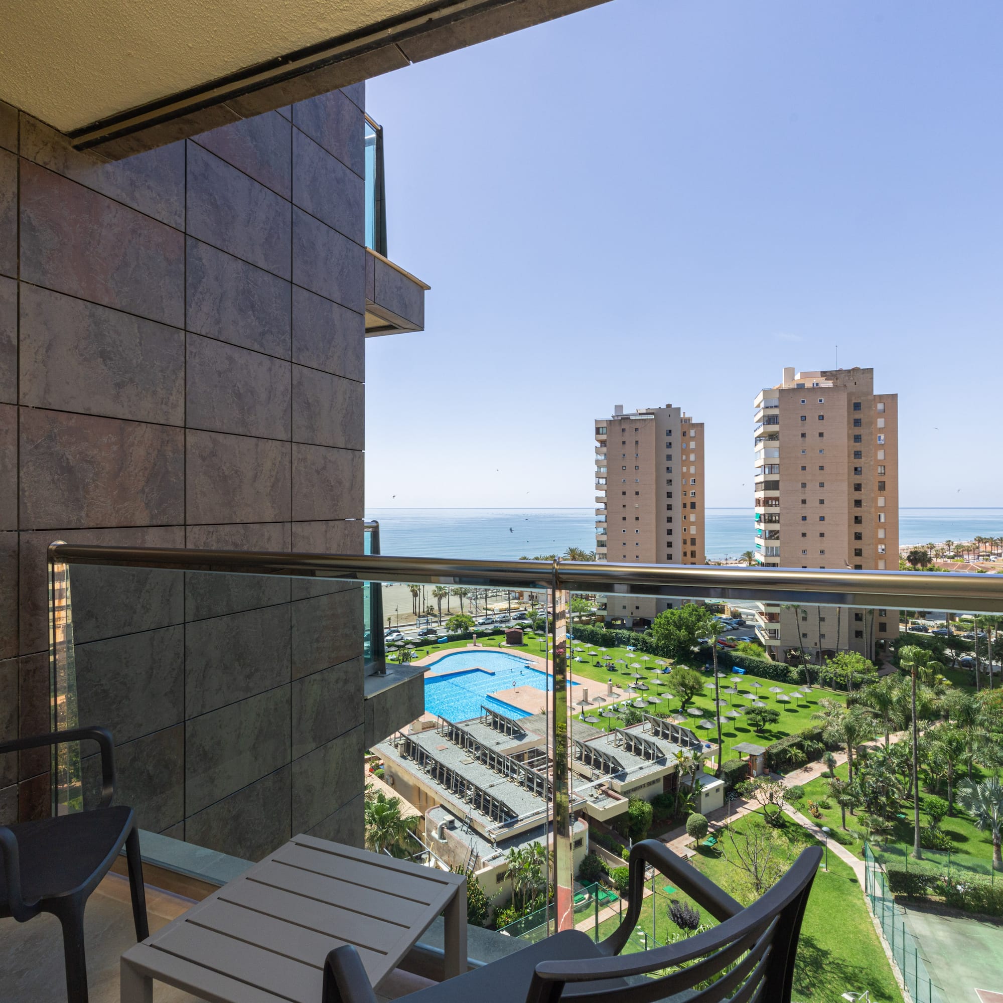 Balcony overlooking sea, resort pool, palm trees, and tall city buildings.