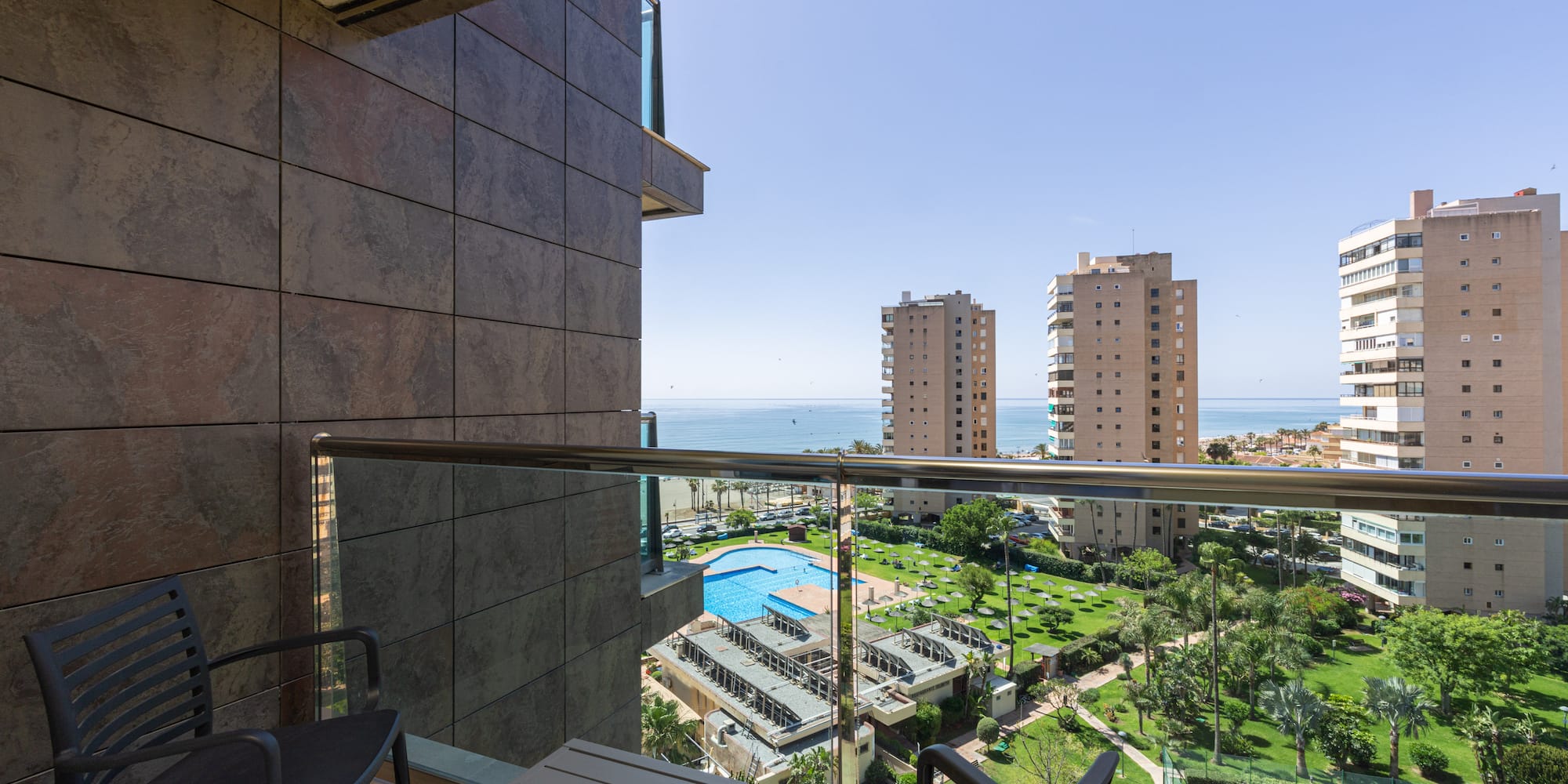 Balcony overlooking sea, resort pool, palm trees, and tall city buildings.