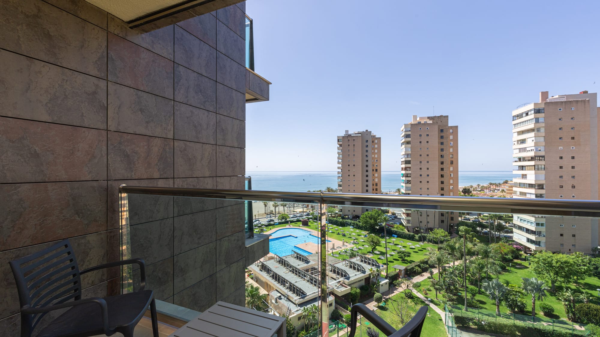 Balcony overlooking sea, resort pool, palm trees, and tall city buildings.