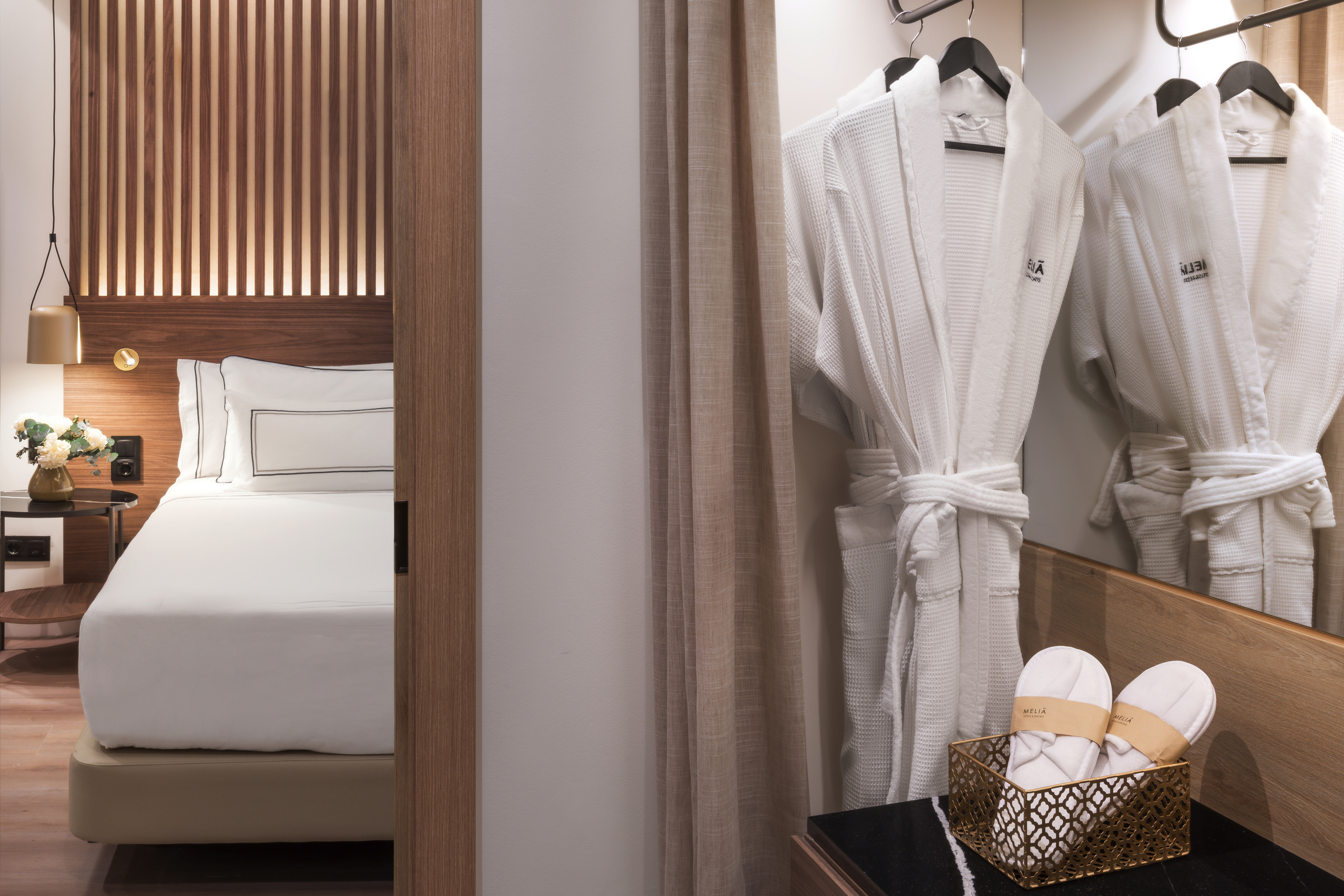 Modern hotel bedroom with white bed, wooden headboard, hanging bathrobes, and slippers.