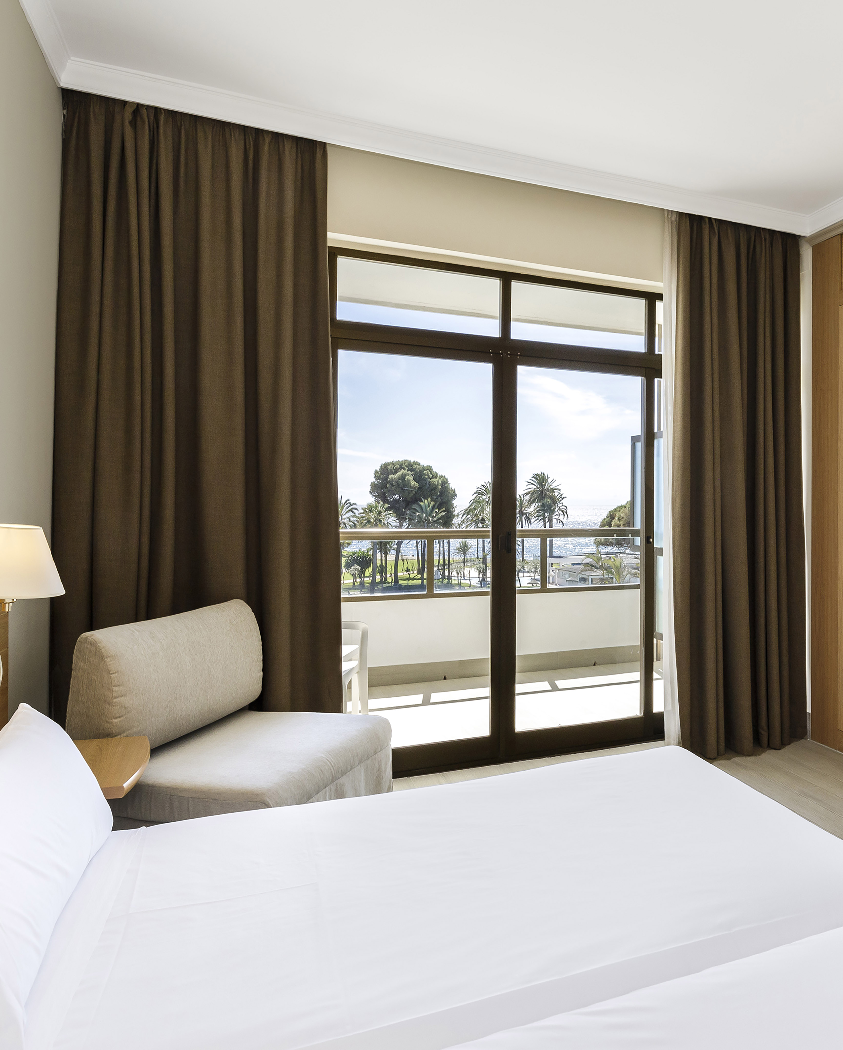 Hotel room with sea view balcony, white bed, and brown curtains.