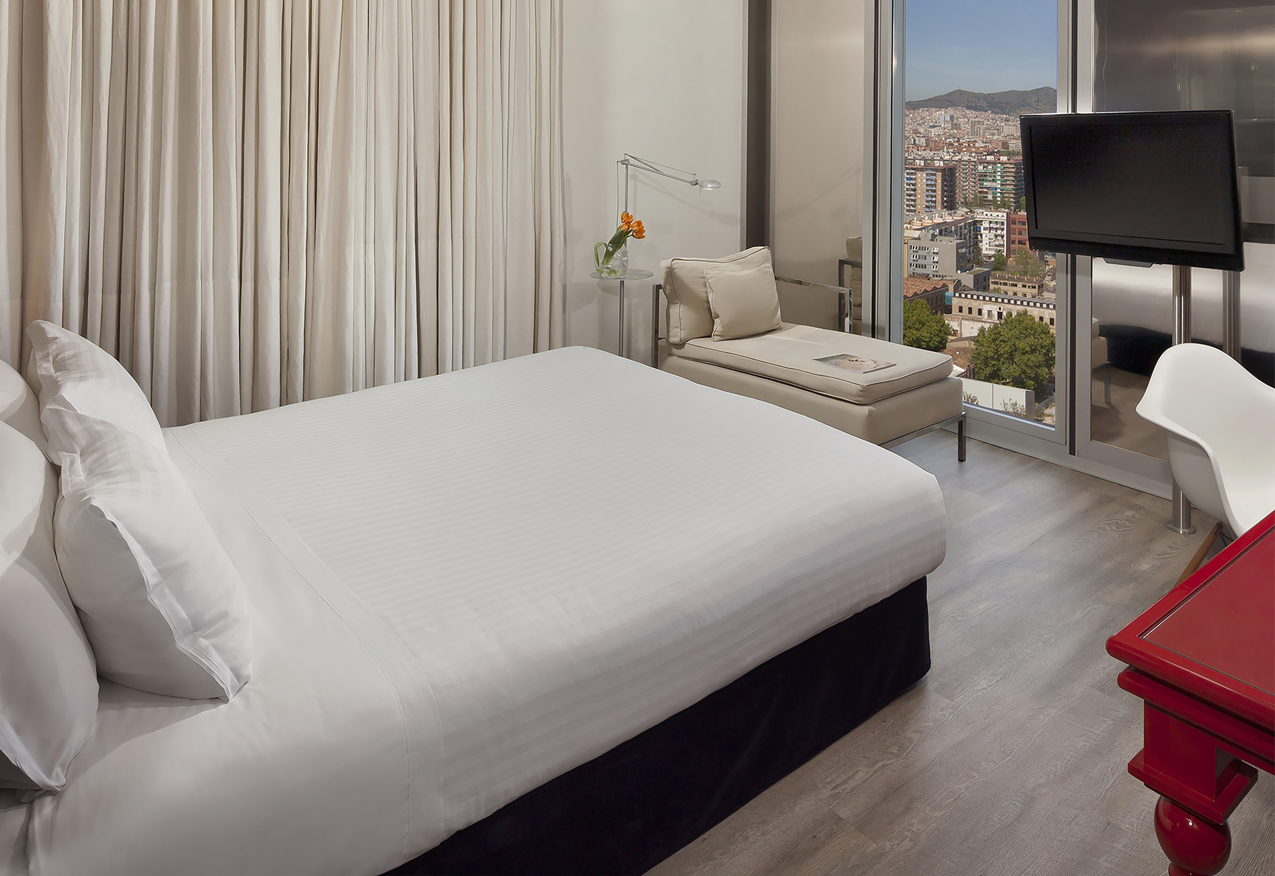 Modern Barcelona hotel room with city view, king bed, chaise lounge, and TV.