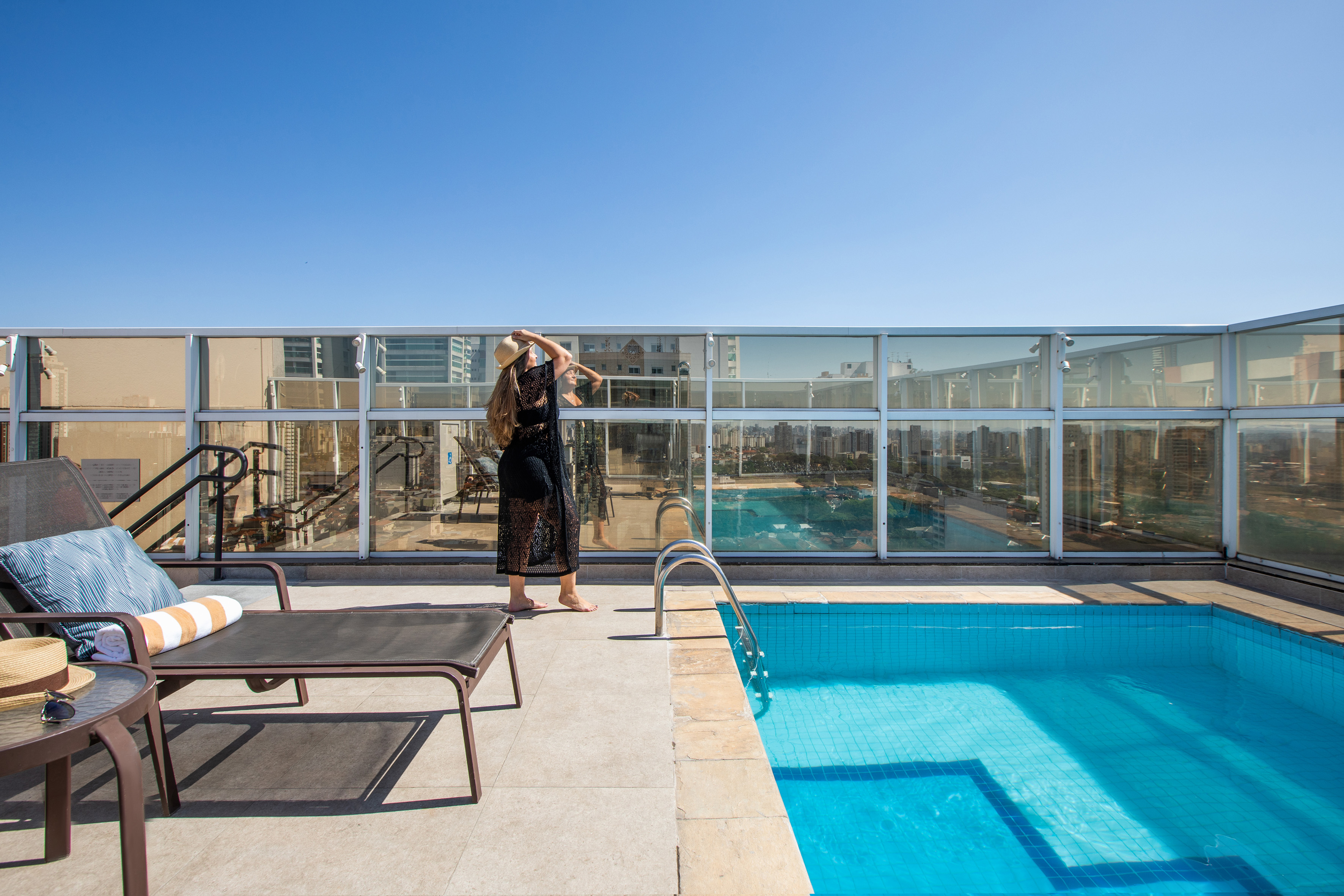 Woman in hat by rooftop swimming pool, city skyline, lounge chair.