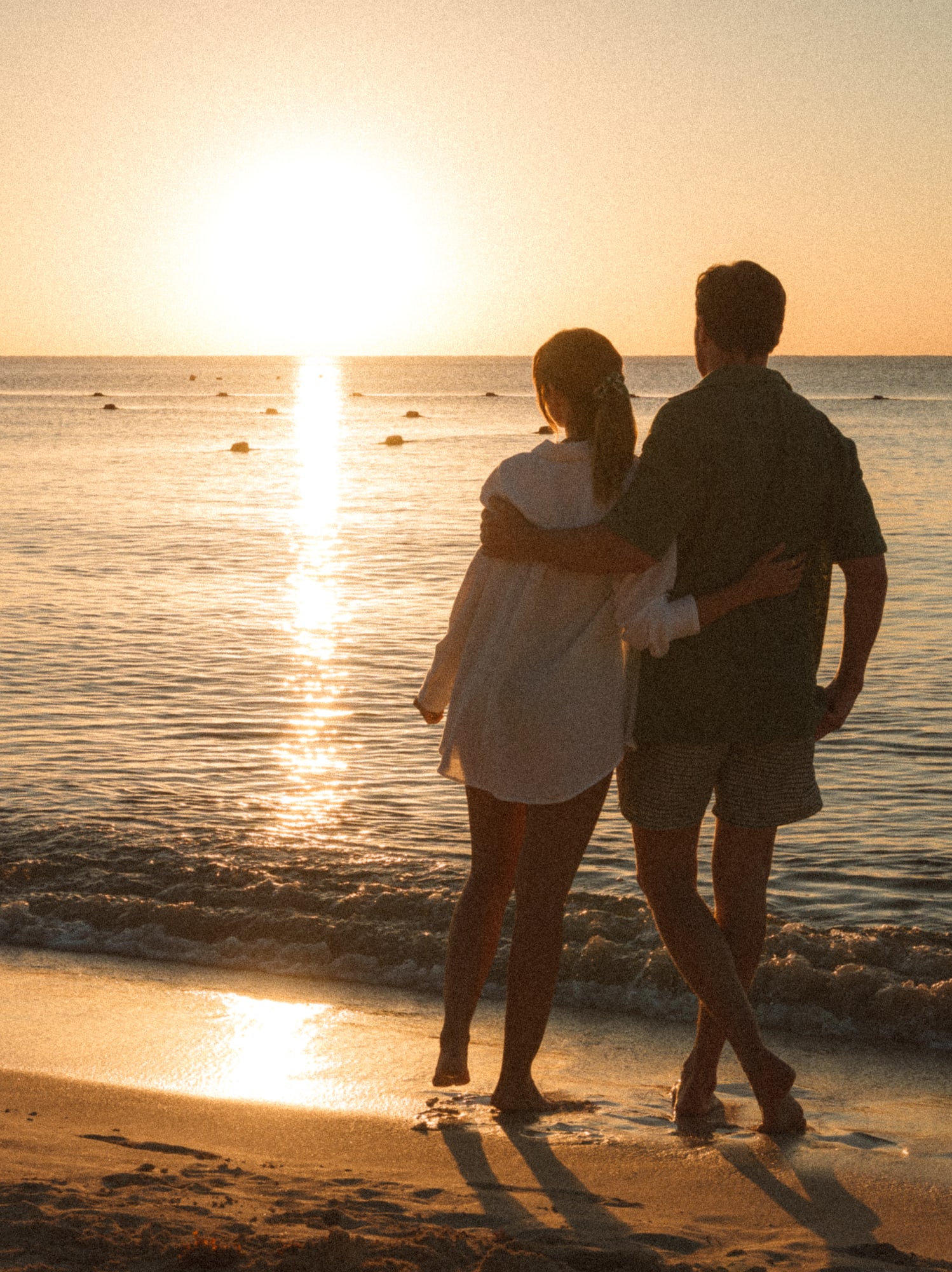 a man and woman standing on a beach looking at the sunset
