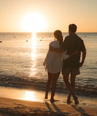 a man and woman standing on a beach looking at the sunset
