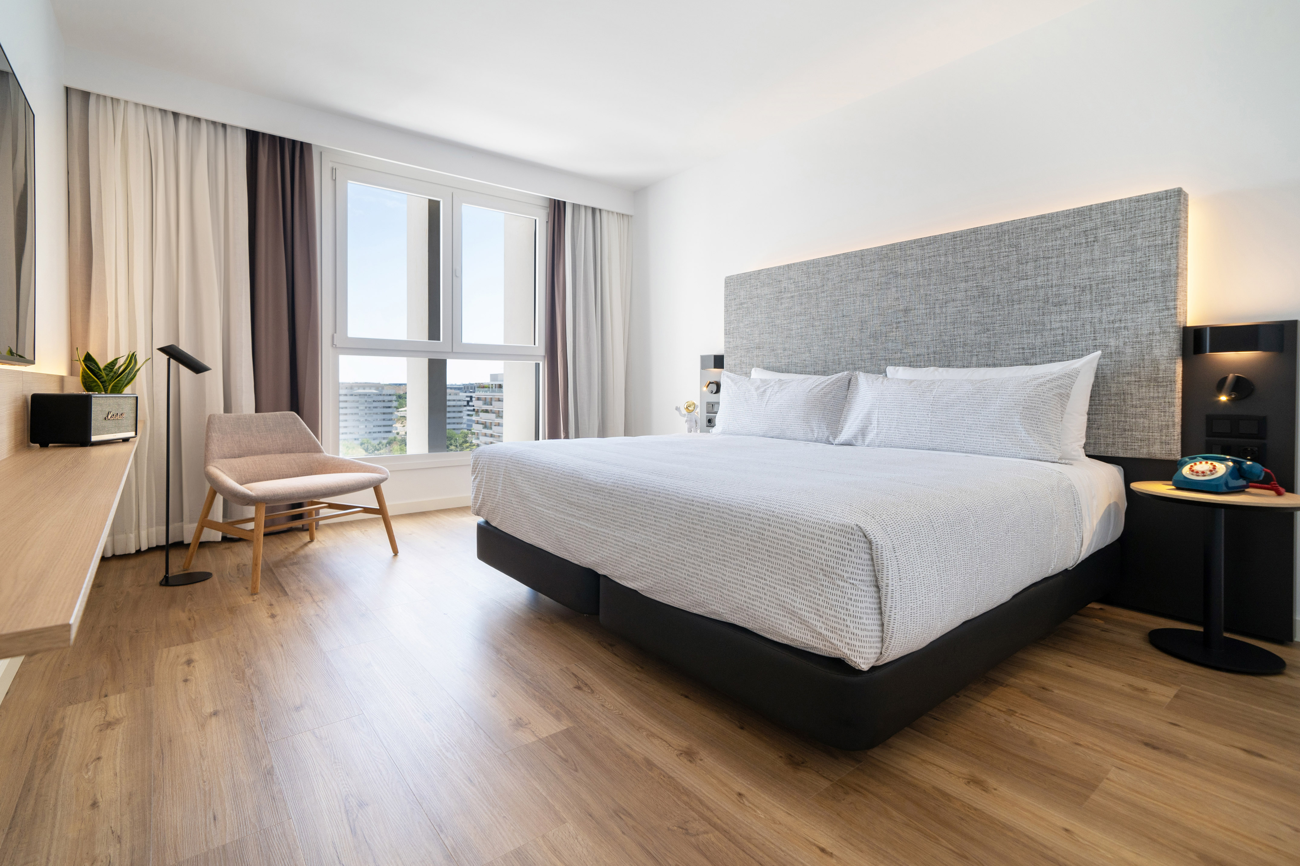Modern hotel bedroom with white bed, hardwood floor, large window, city view.