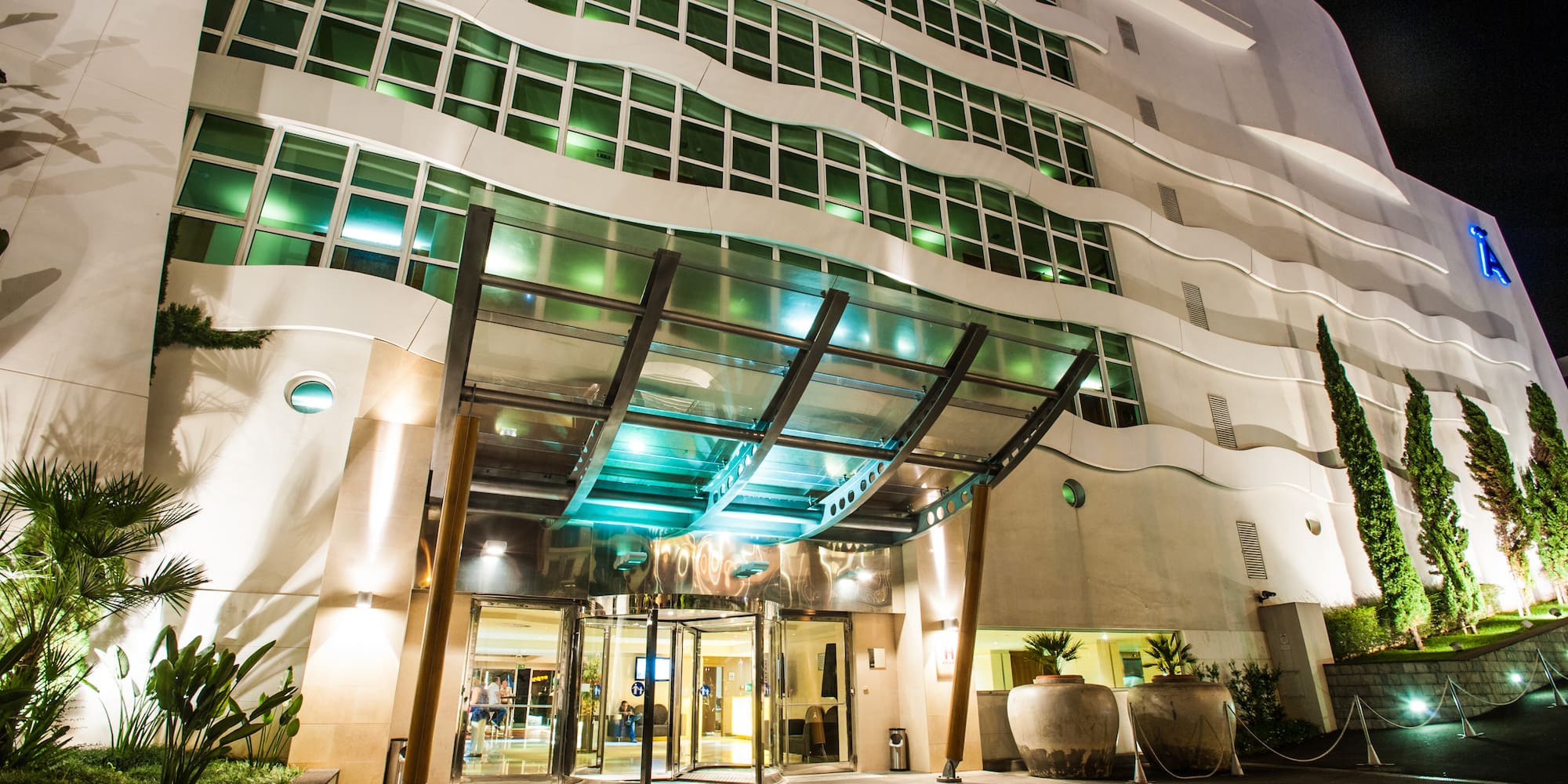Modern hotel facade at night, illuminated entrance, wavy design, green windows.