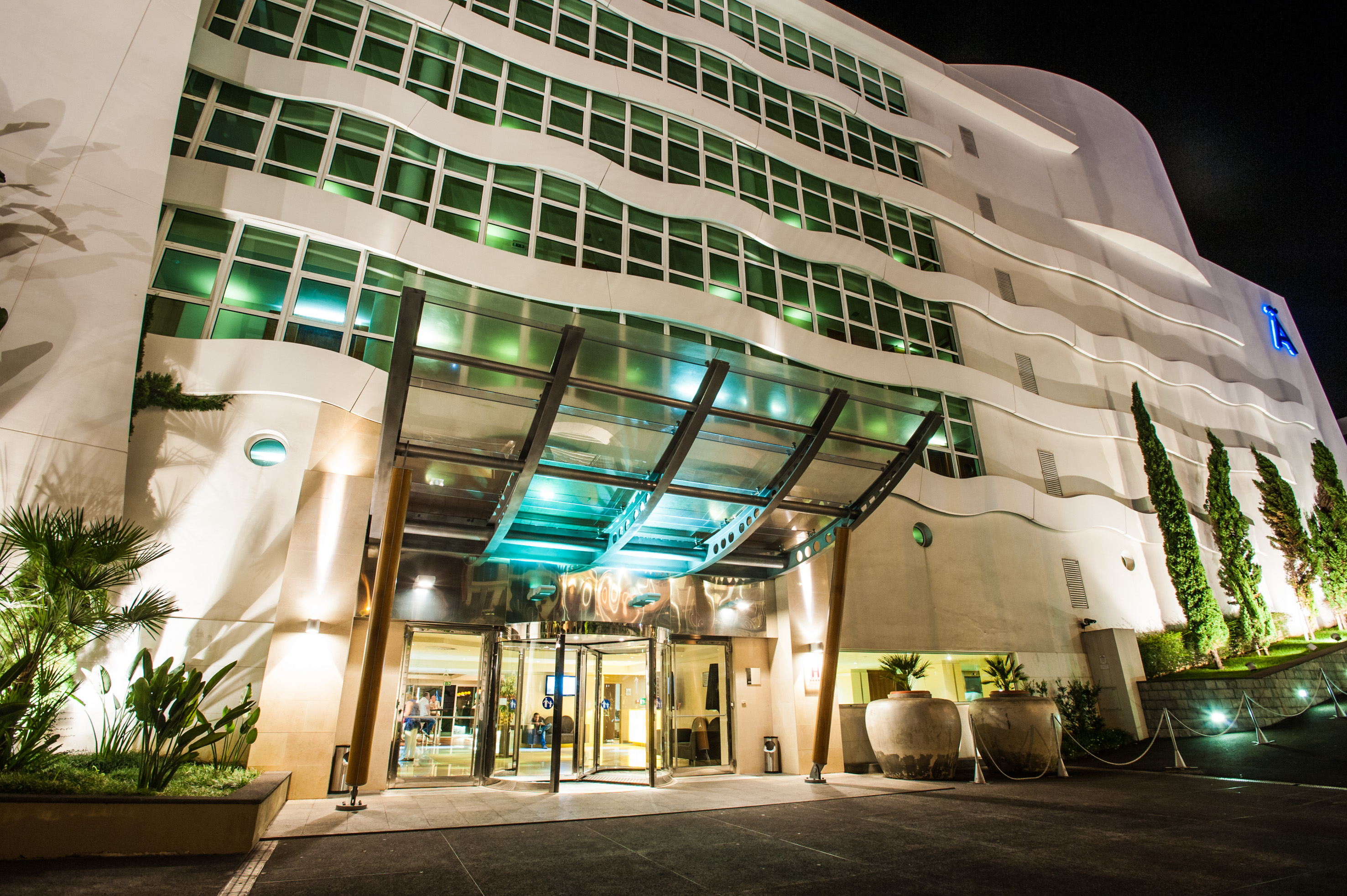 Modern hotel facade at night, illuminated entrance, wavy design, green windows.