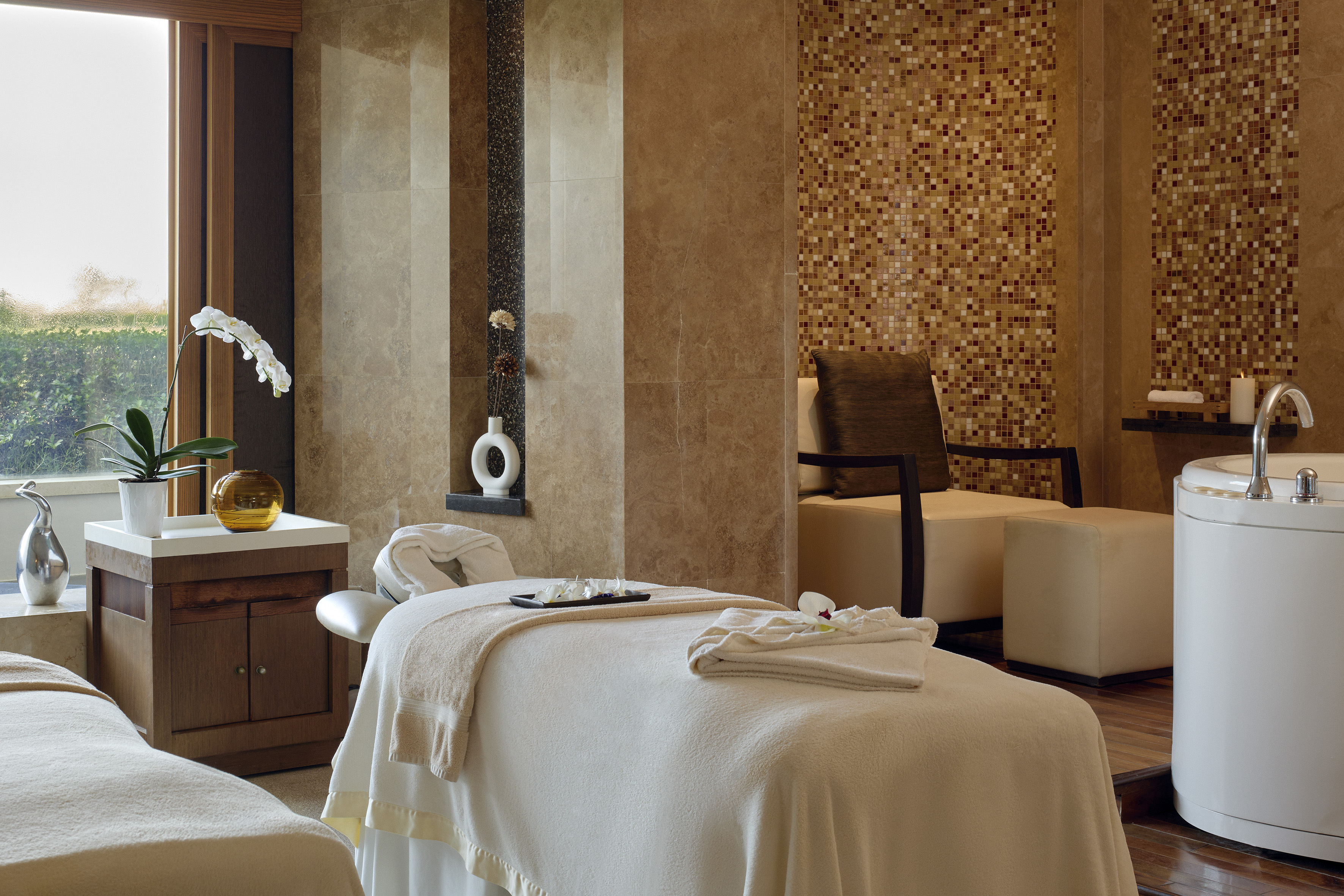 Luxury spa treatment room with massage beds, window view, orchid, and bathtub.