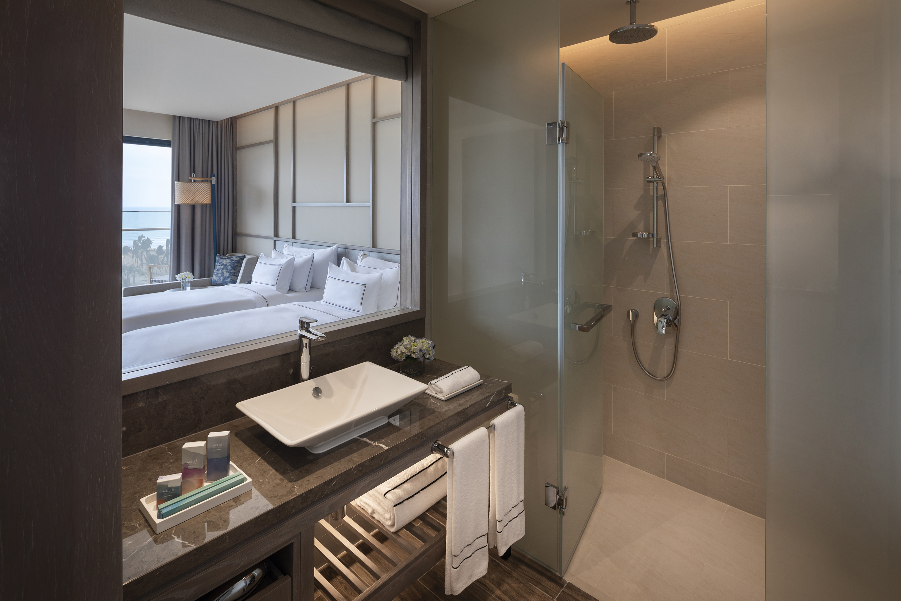 Modern hotel bathroom, vanity, glass shower, reflecting twin beds, ocean view.