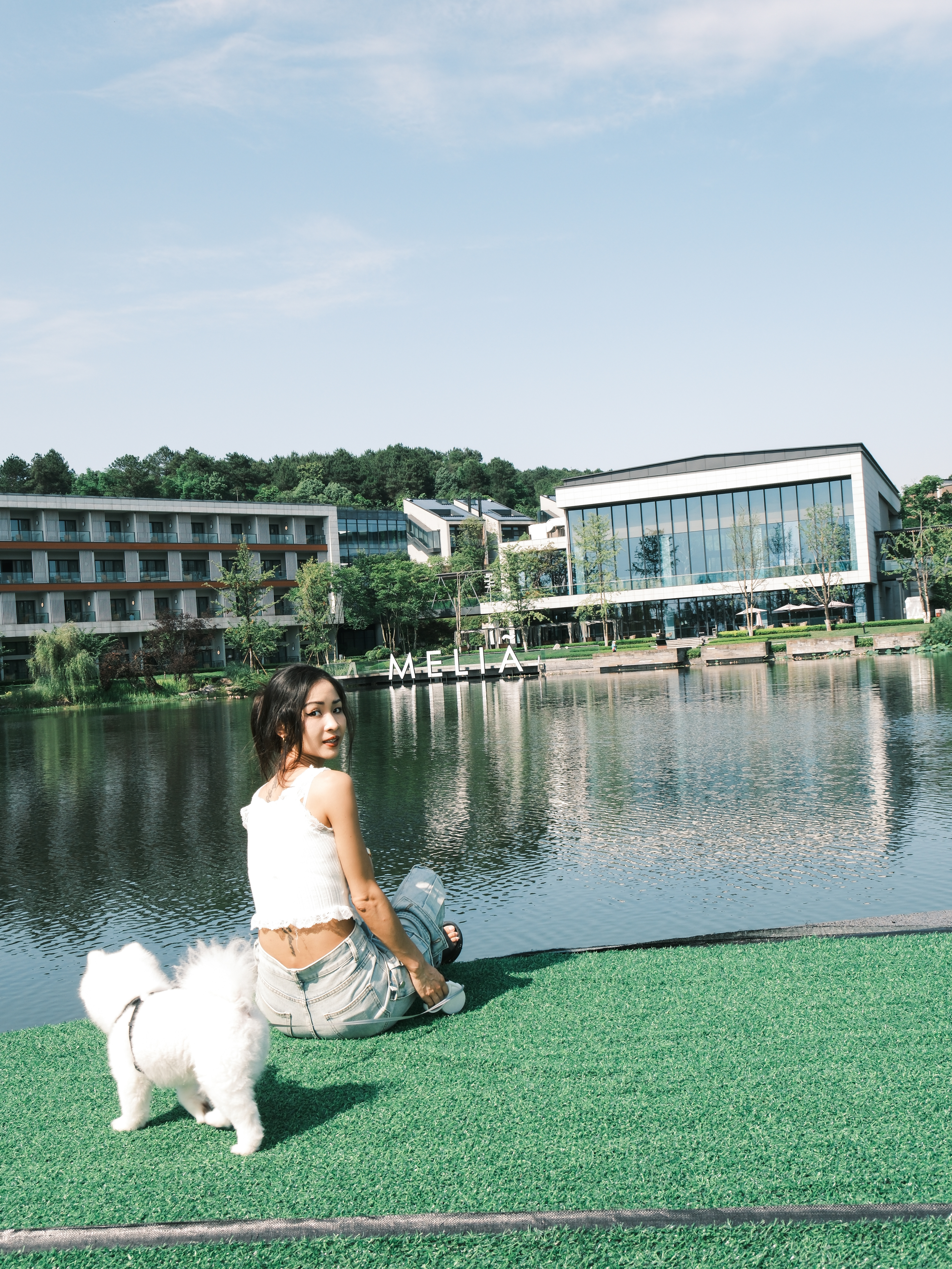 Woman and white dog sit by a lake with Melia hotel buildings.