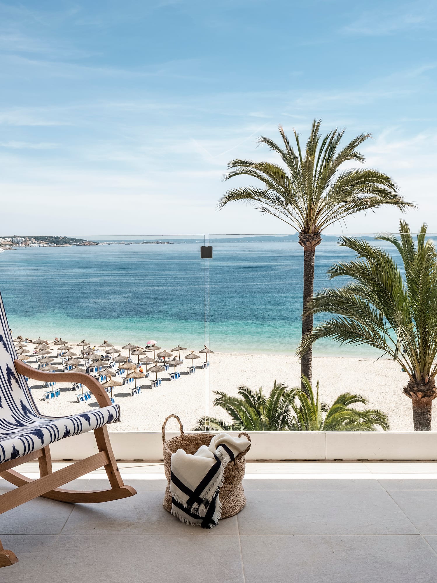Resort balcony with rocking chair, beach, palm trees, and clear blue sea view.