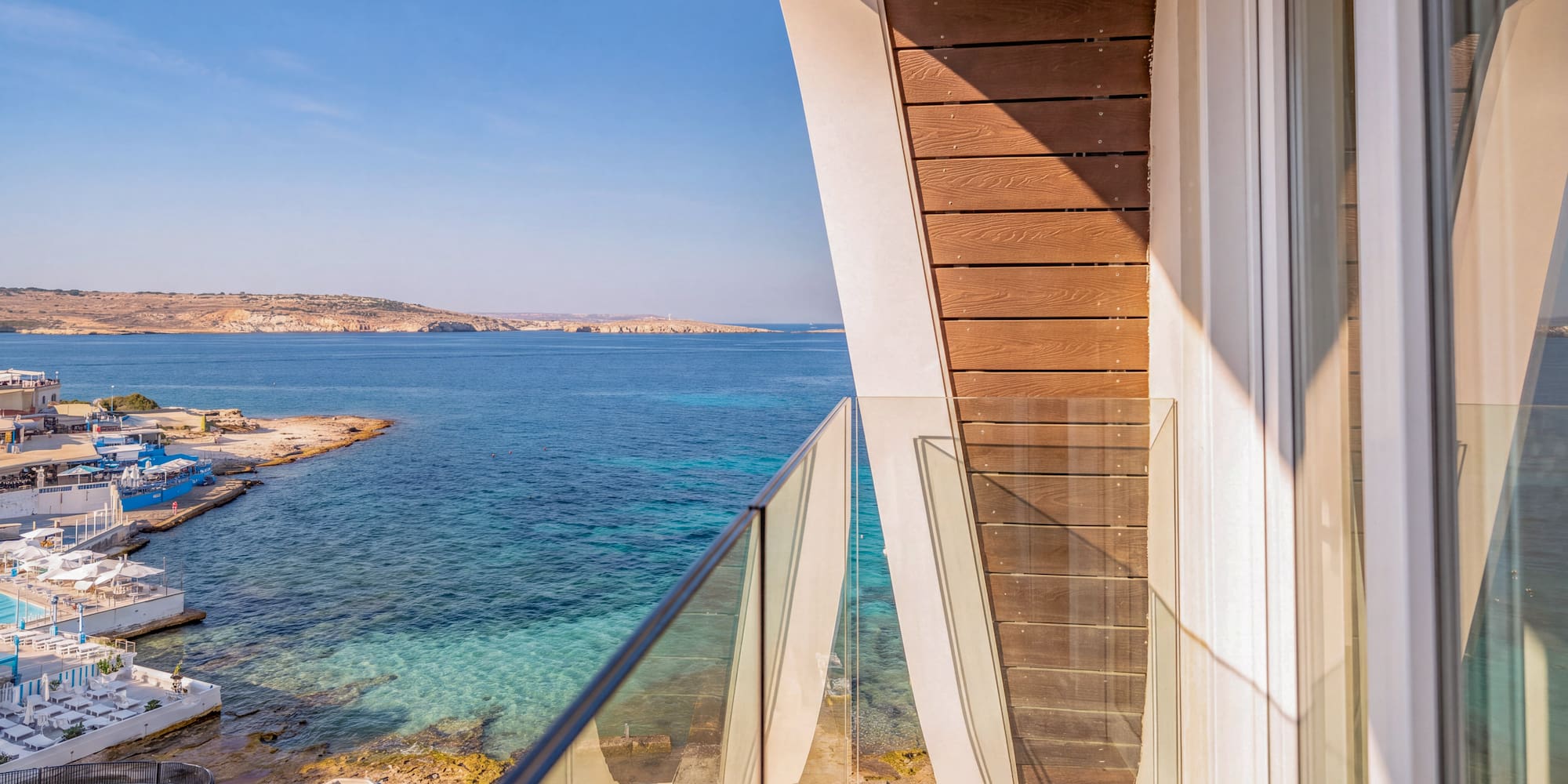 Modern balcony with glass railing overlooking clear blue sea and coastal resort.
