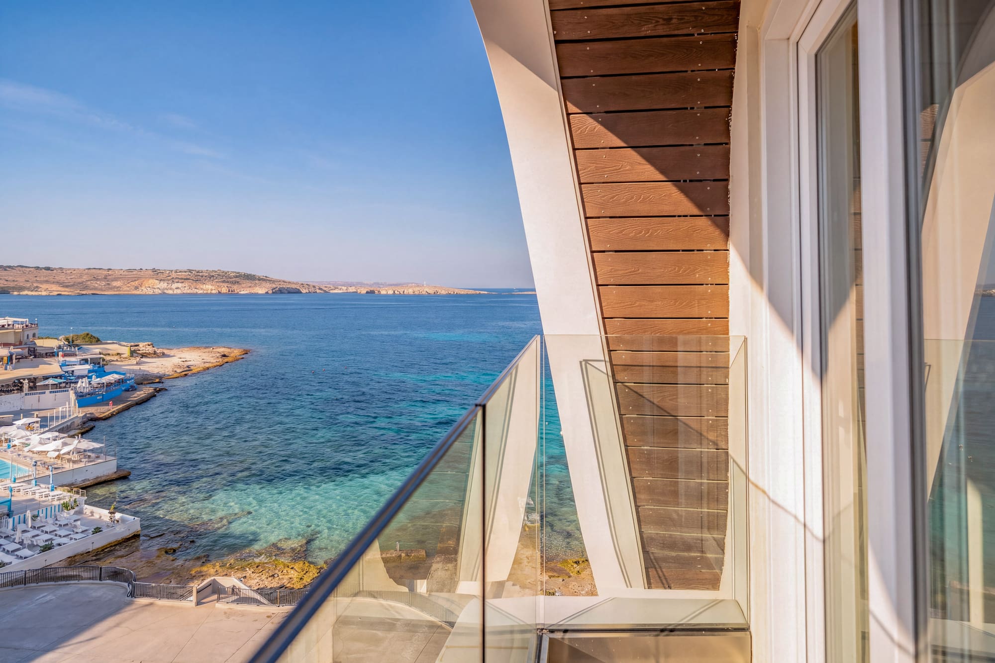 Modern balcony with glass railing overlooking clear blue sea and coastal resort.