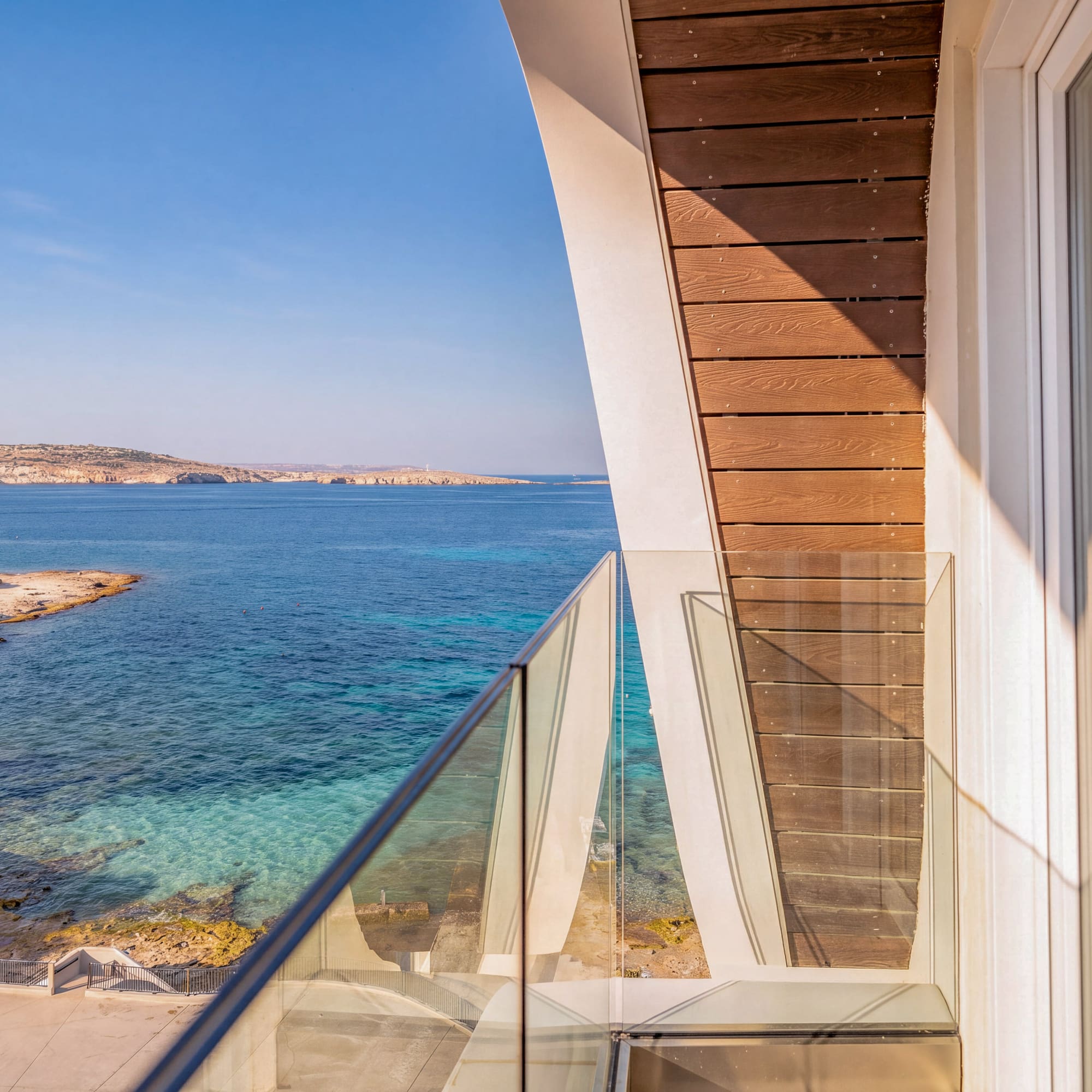 Modern balcony with glass railing overlooking clear blue sea and coastal resort.