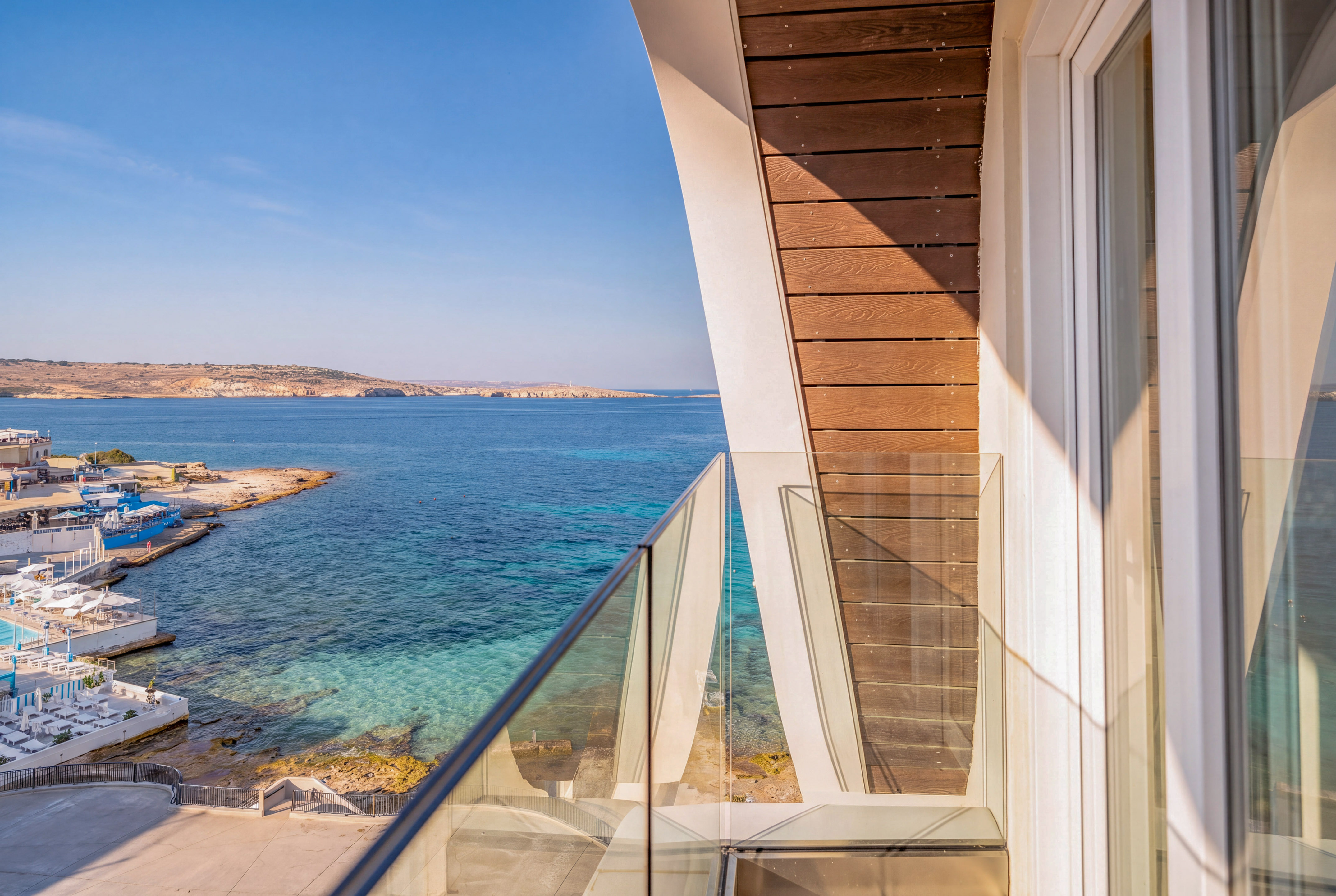 Modern balcony with glass railing overlooking clear blue sea and coastal resort.