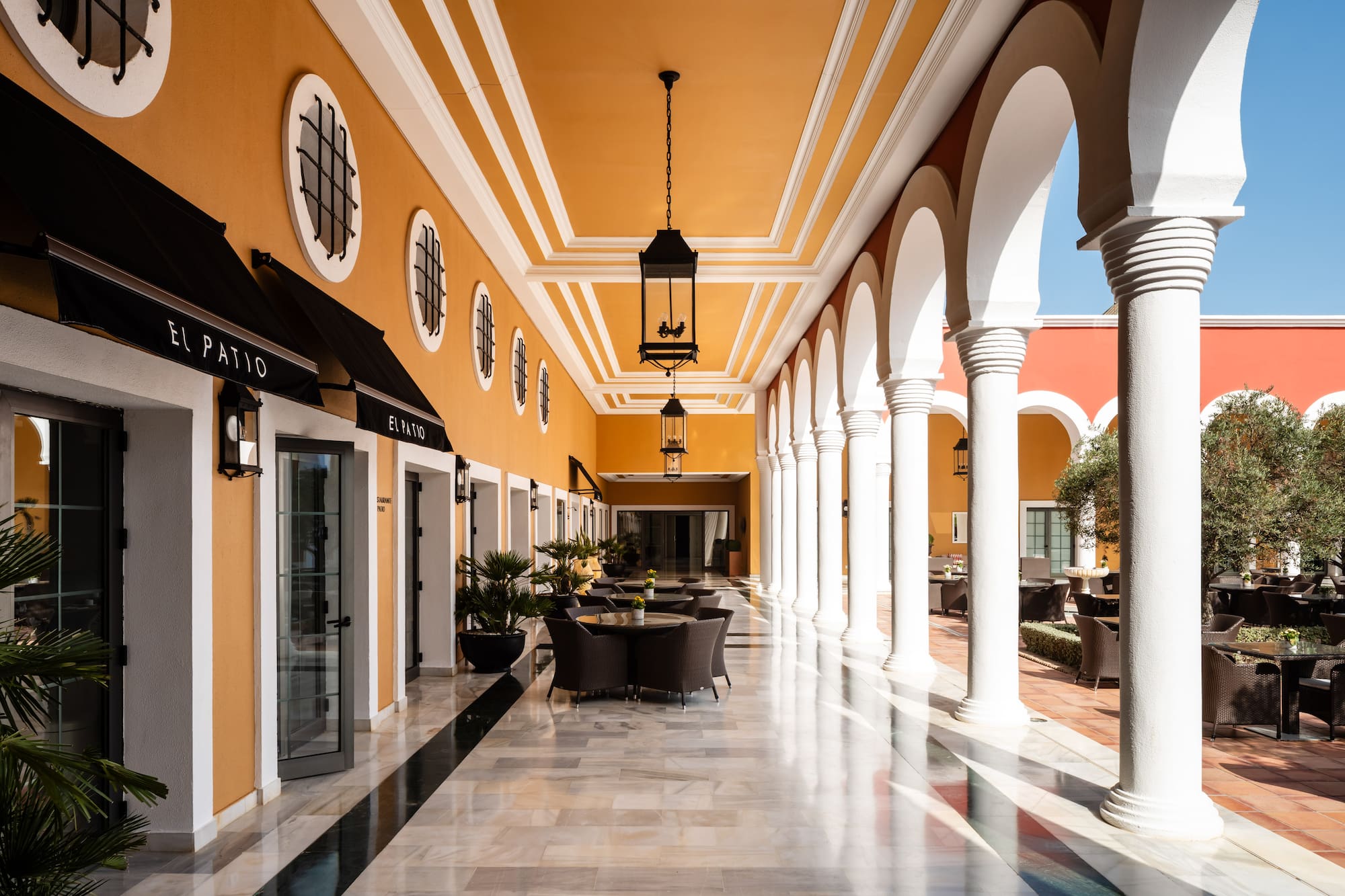 El Patio hotel walkway with orange walls, white arches, marble floor & outdoor cafe.