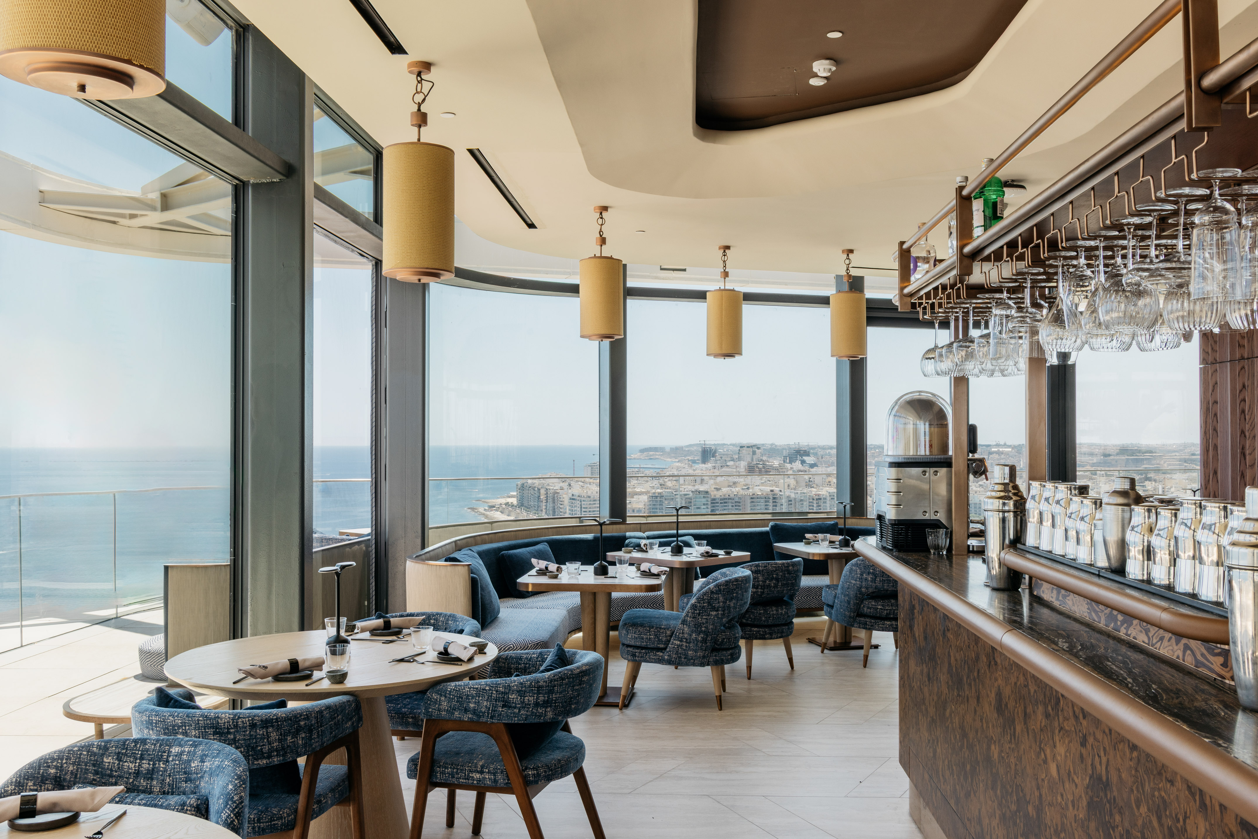 Modern restaurant with panoramic sea & city views, stylish bar, blue seating.