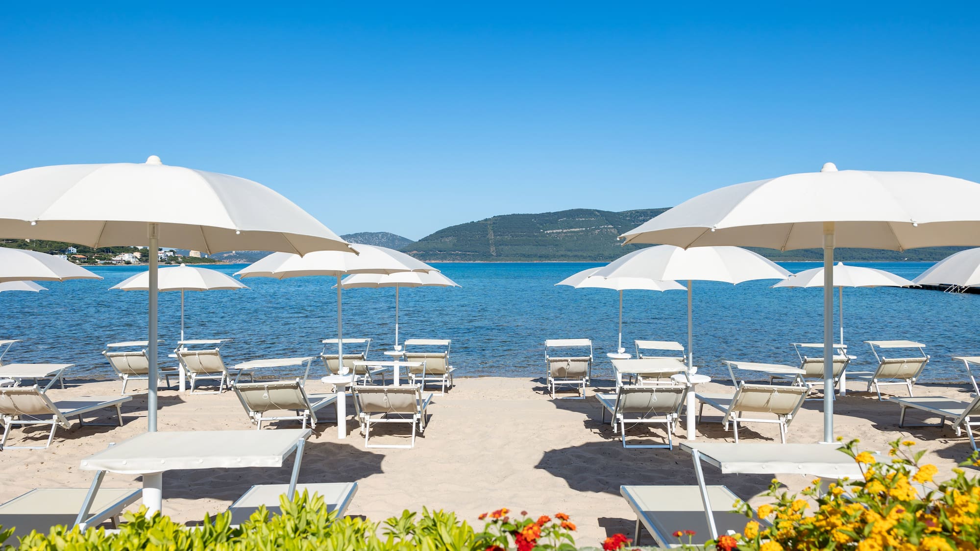 Sunny beach with white umbrellas, lounge chairs, blue sea, and colorful flowers.