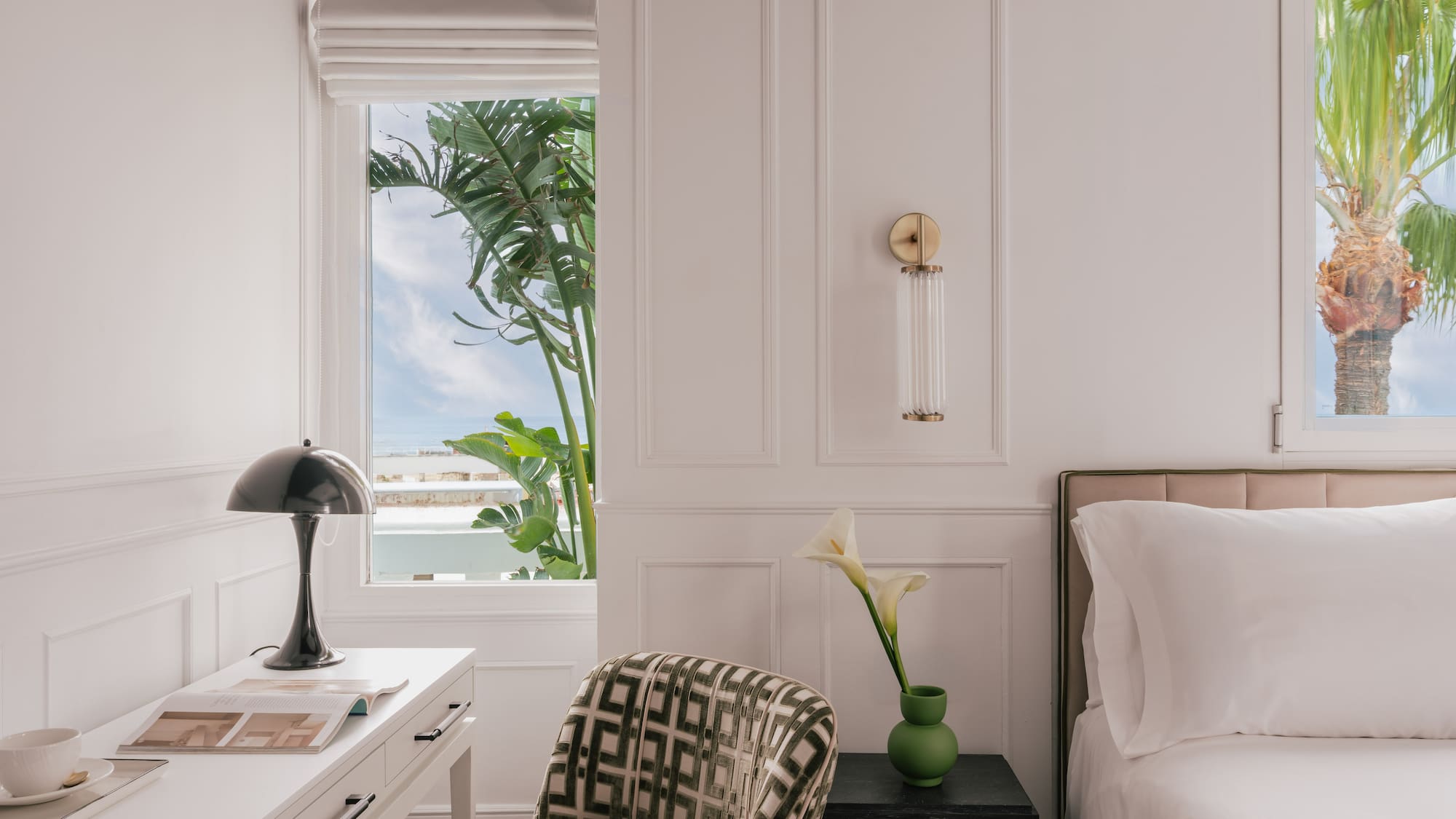 Modern white bedroom with desk, patterned chair, and tropical palm view.