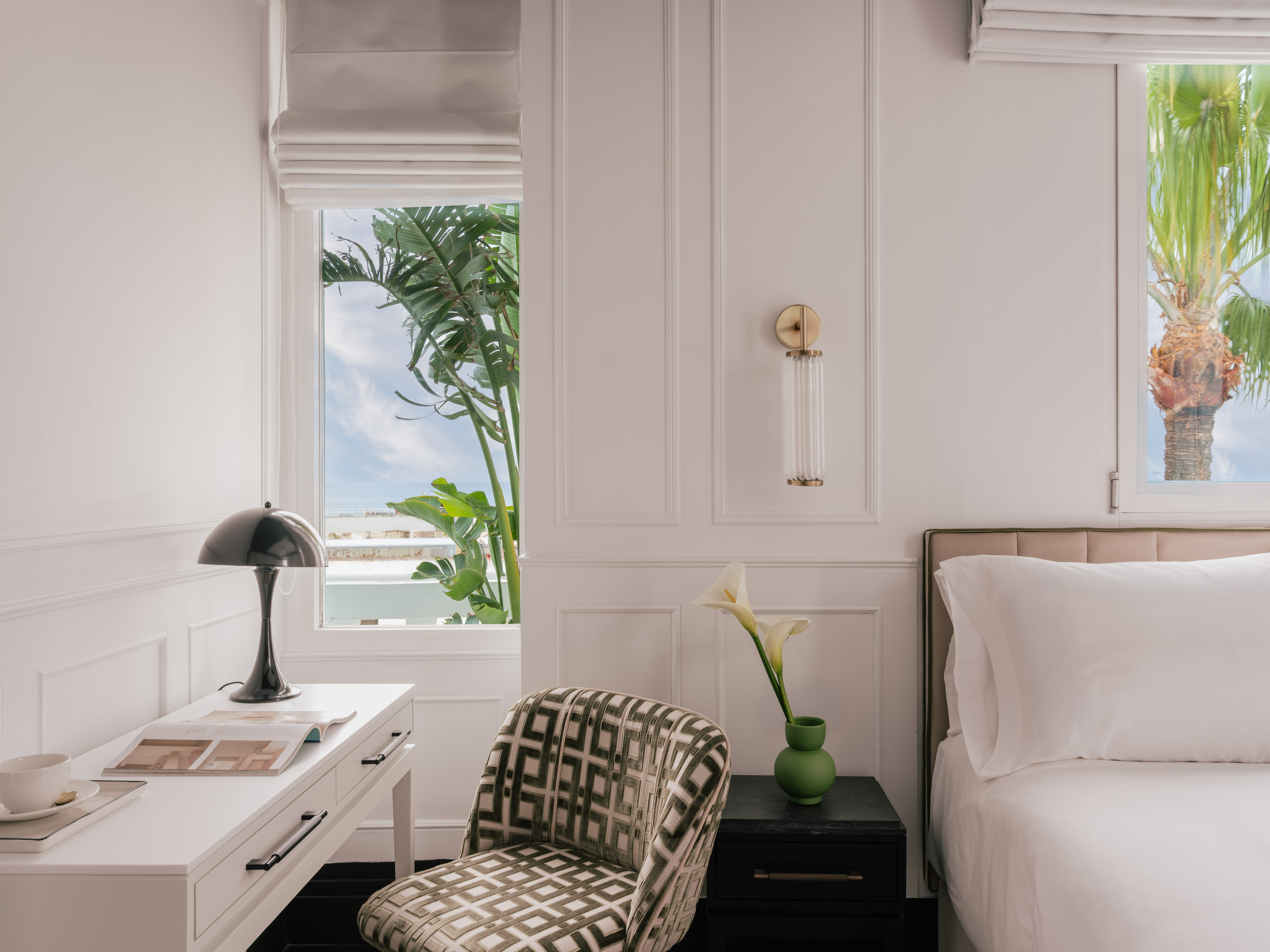 Modern bedroom: white decor, desk, bed, and windows with tropical palm trees.