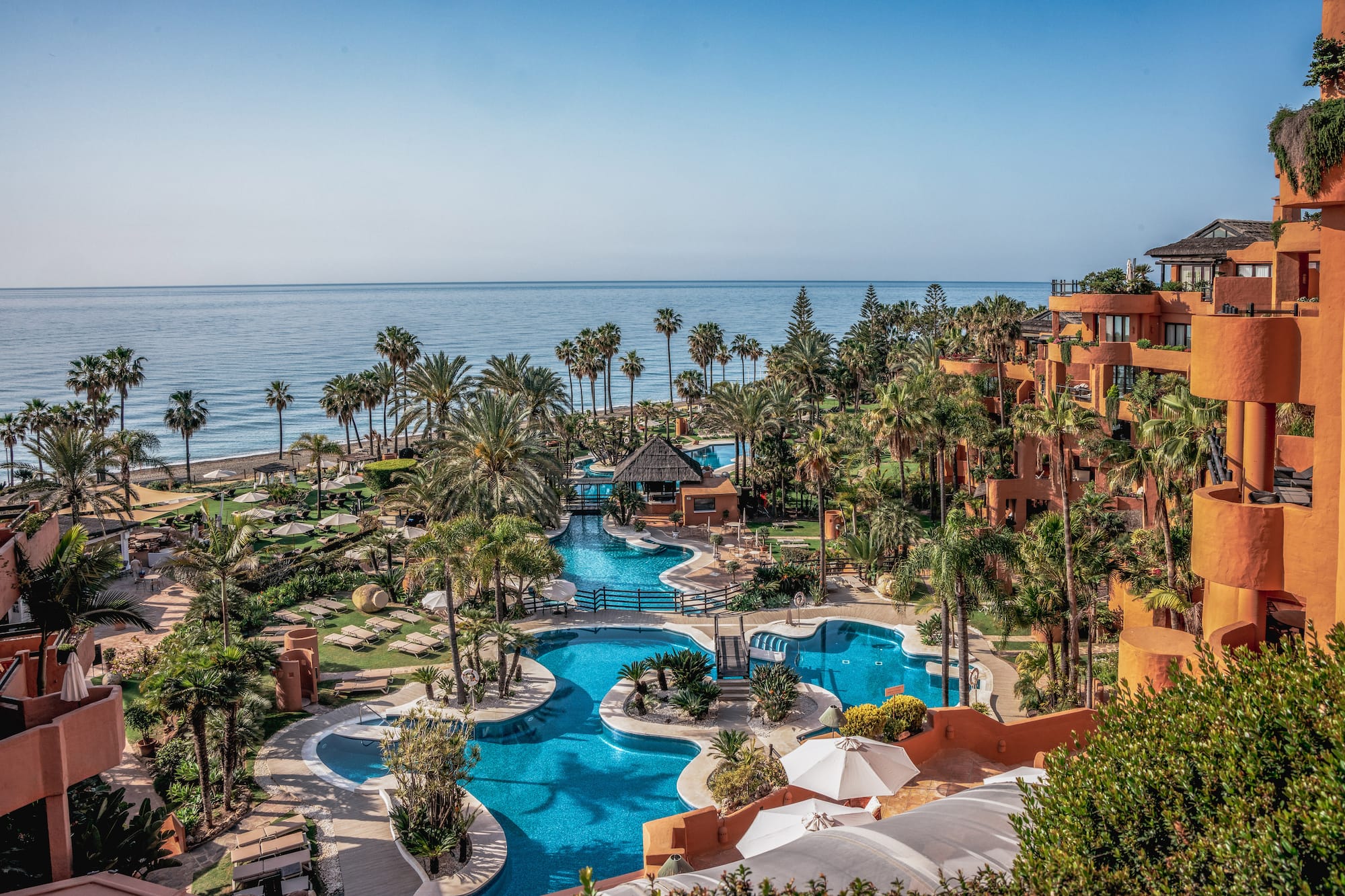 Oceanfront resort with winding blue pools, palm trees, and terracotta hotel buildings.