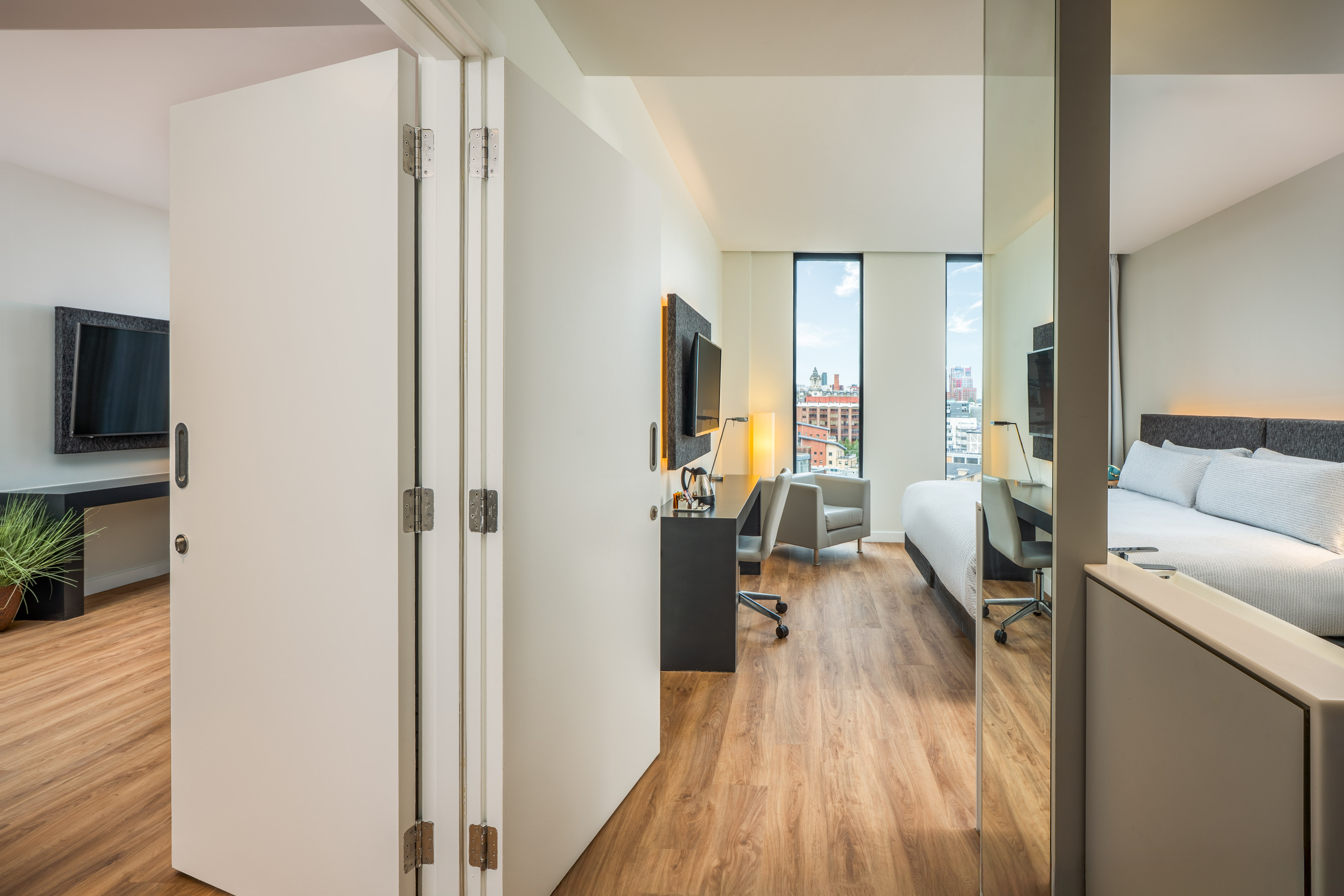 Contemporary hotel room with folding door, wood floor, bed, desk, city view.