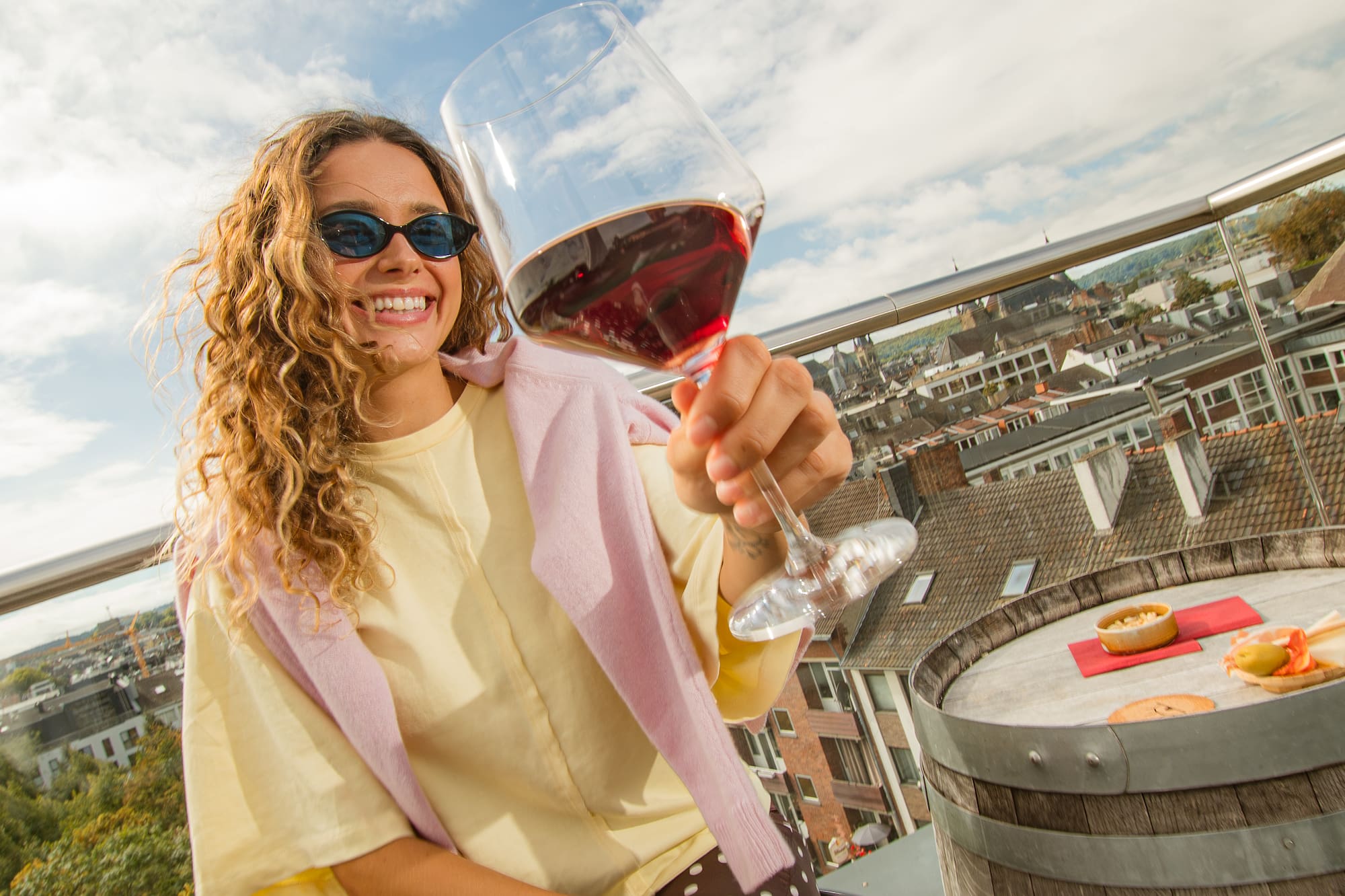 Smiling woman enjoying red wine on sunny rooftop terrace with city view.