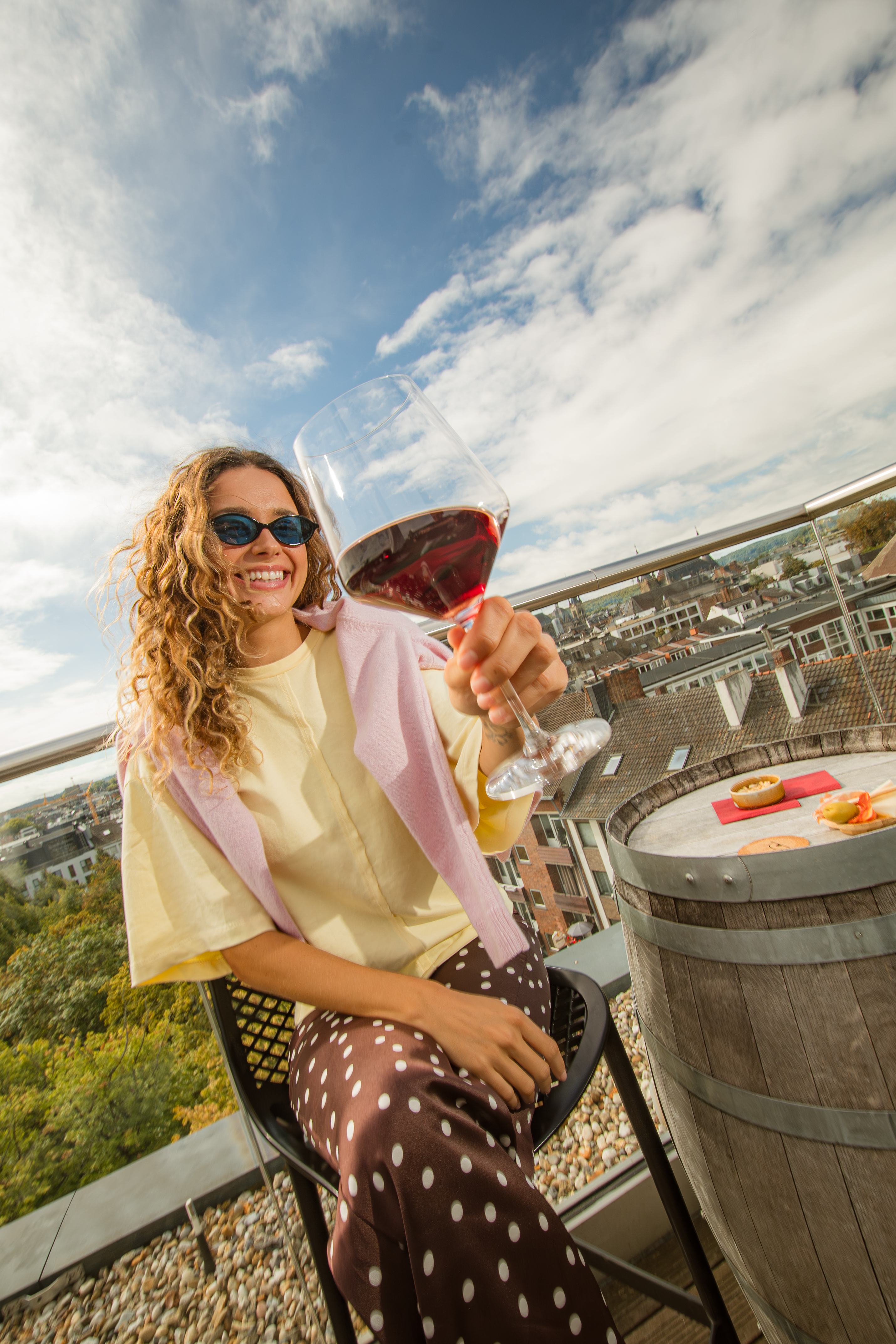 Smiling woman enjoying red wine on sunny rooftop terrace with city view.