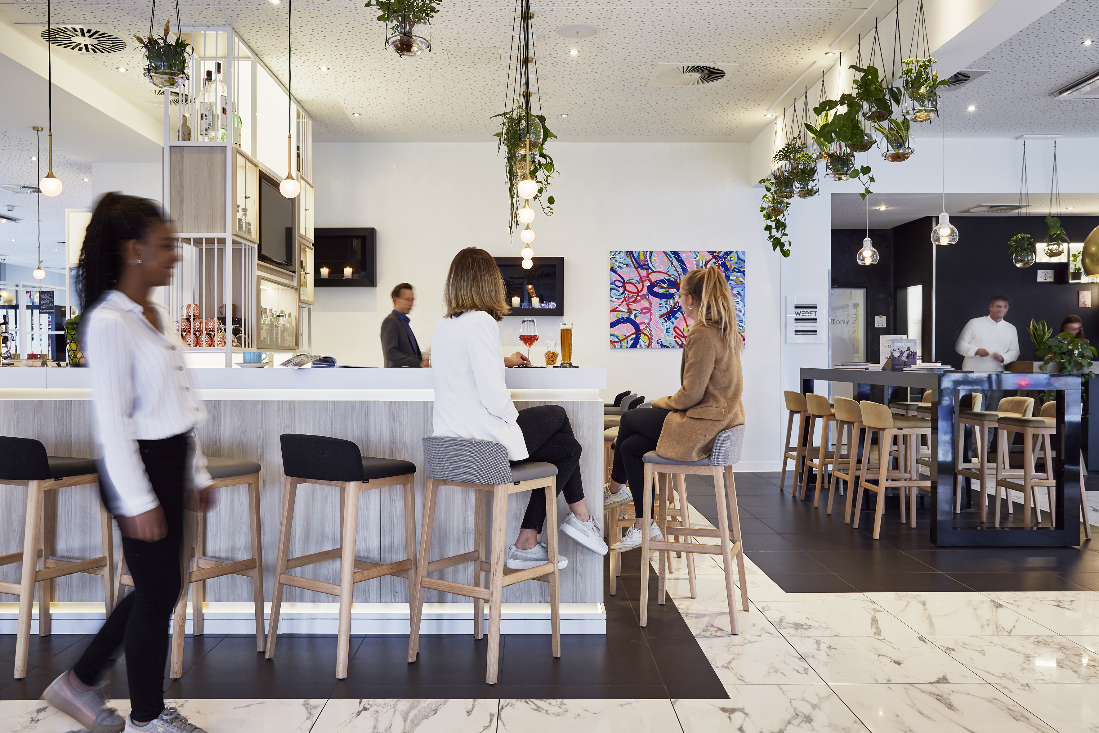 Modern hotel bar lounge with guests, bar counter, stools, and hanging plants.