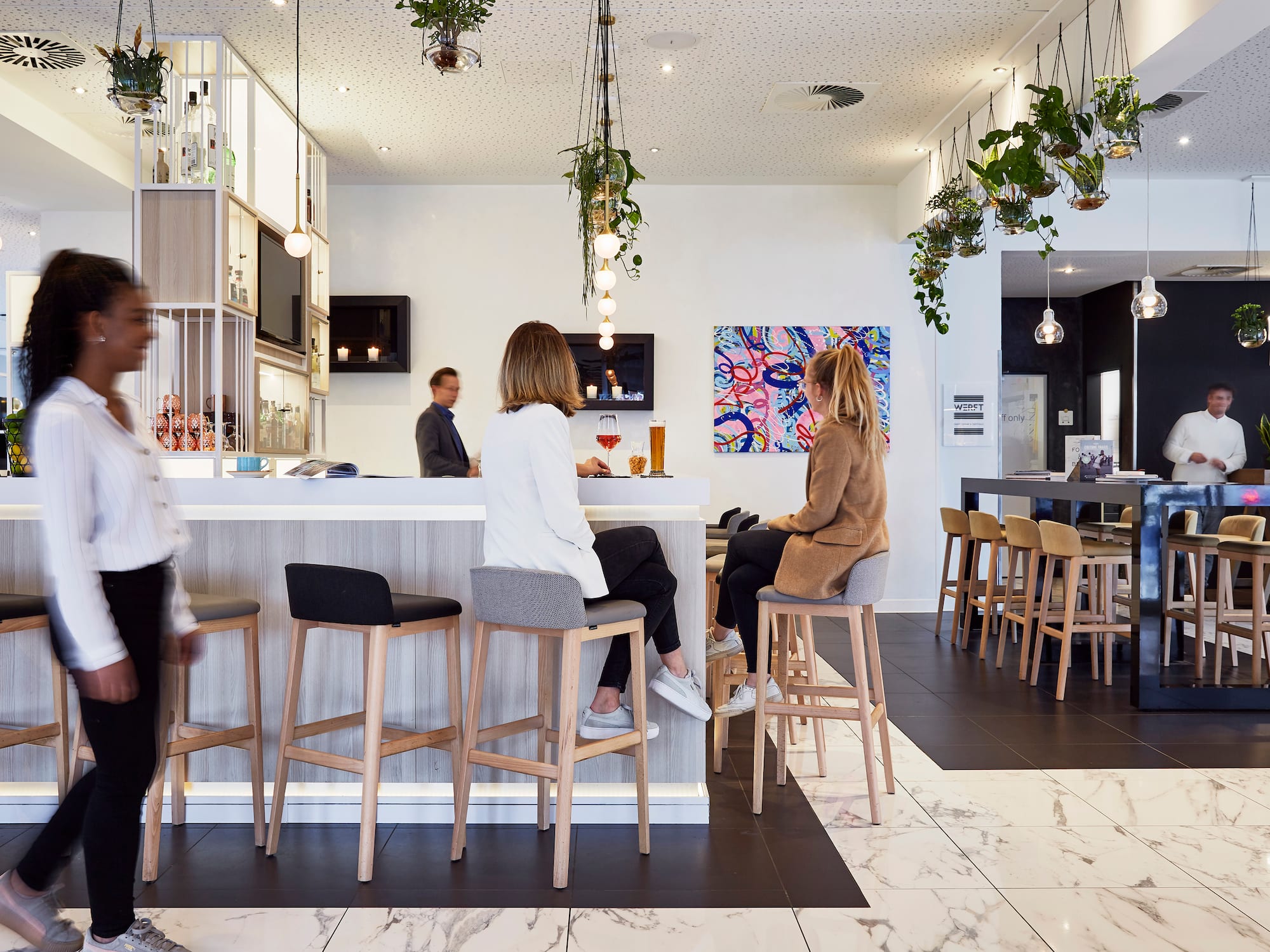 Modern hotel bar lounge with guests, bar counter, stools, and hanging plants.