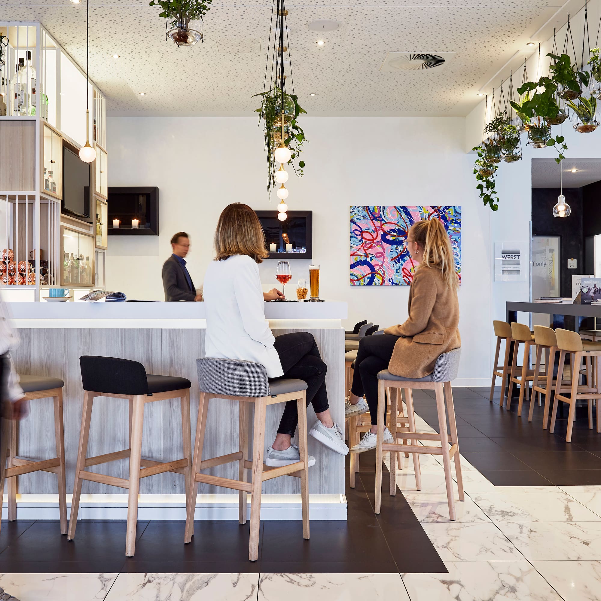 Modern hotel bar lounge with guests, bar counter, stools, and hanging plants.