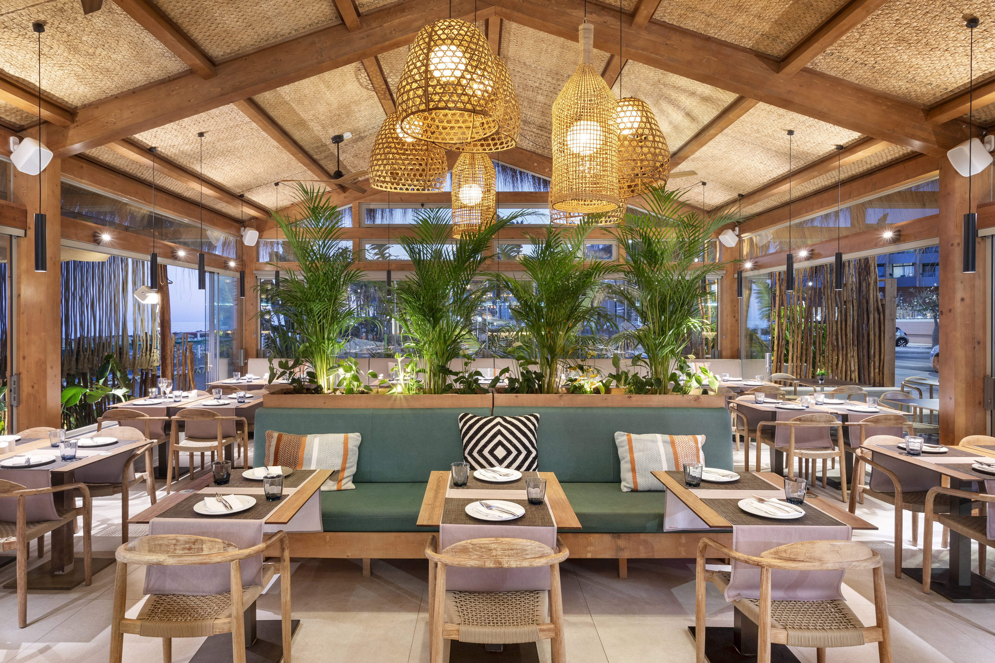 Tropical modern restaurant interior with rattan lights, green seating, and palm plants.
