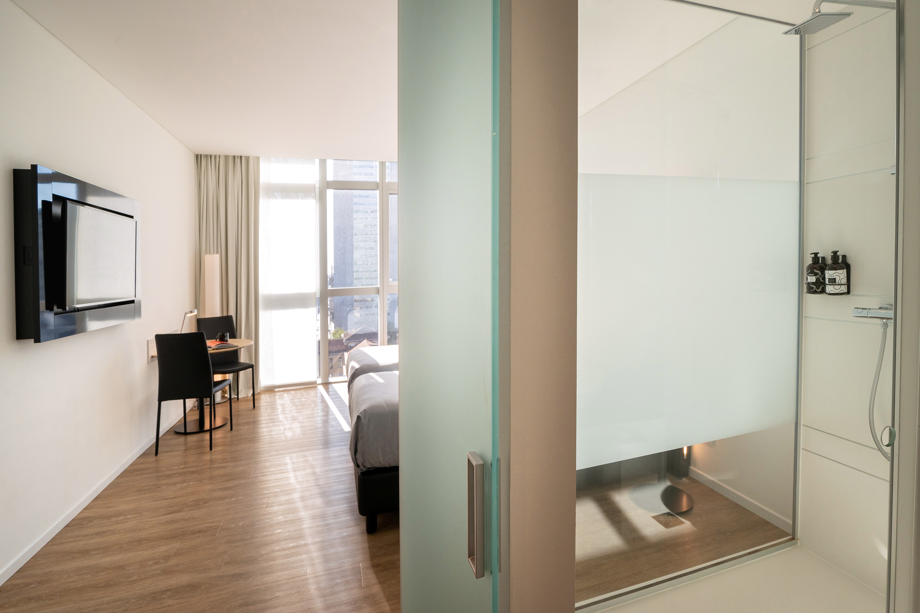 Bright modern hotel room with bed, desk, city view, and glass shower.