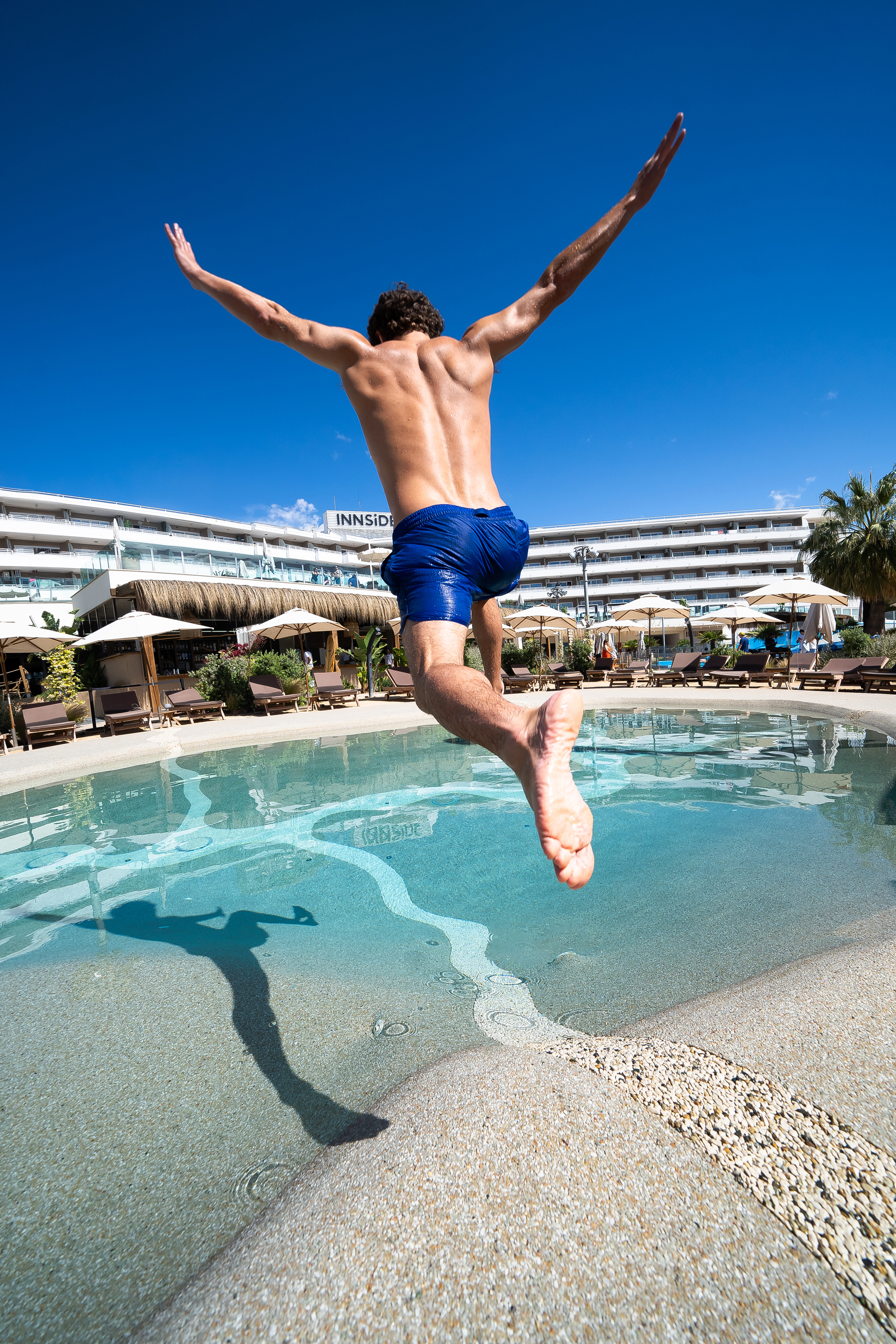 Active man jumping into resort pool under a bright blue sky.