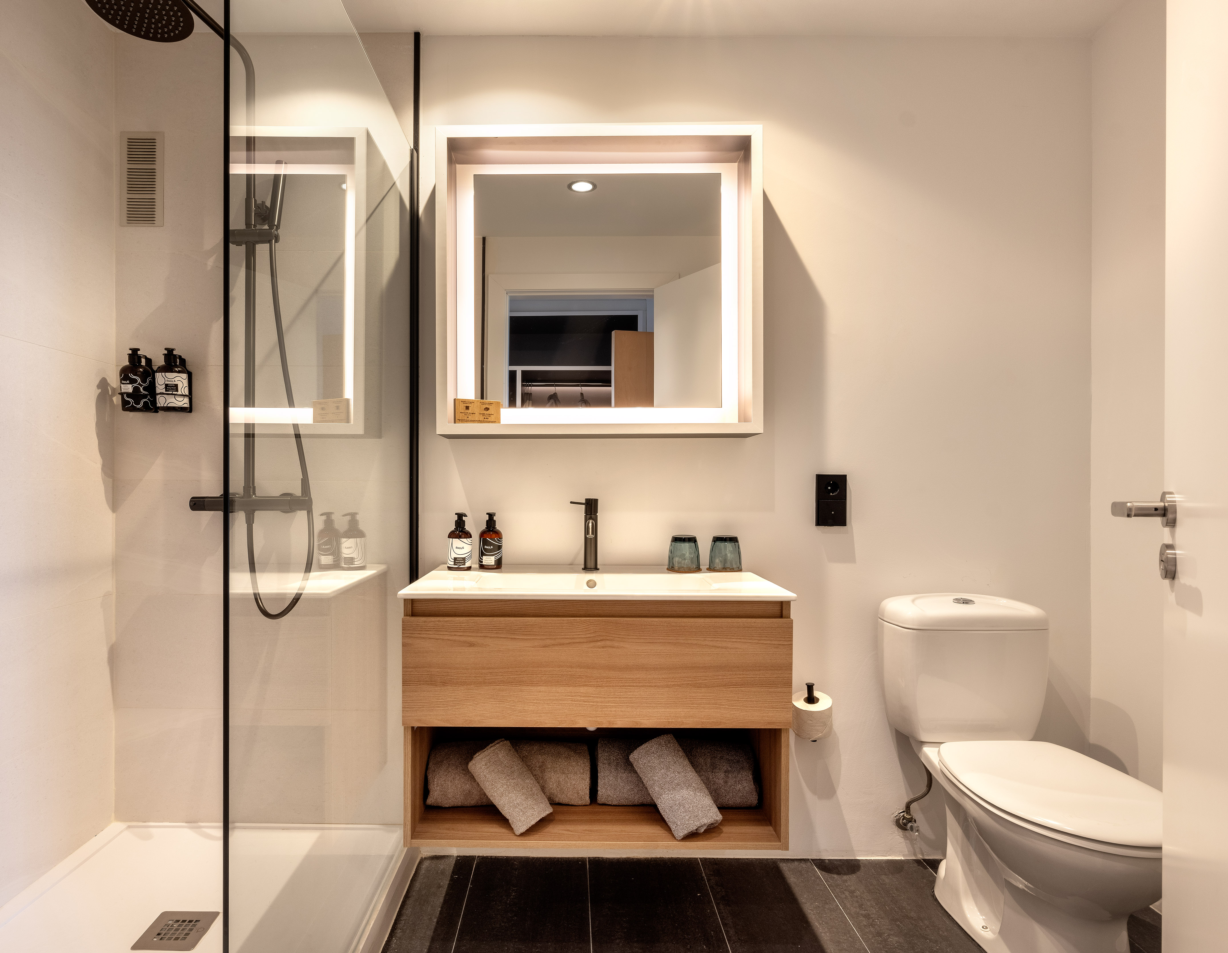 Modern bathroom with walk-in shower, wooden vanity, backlit mirror, and toilet.