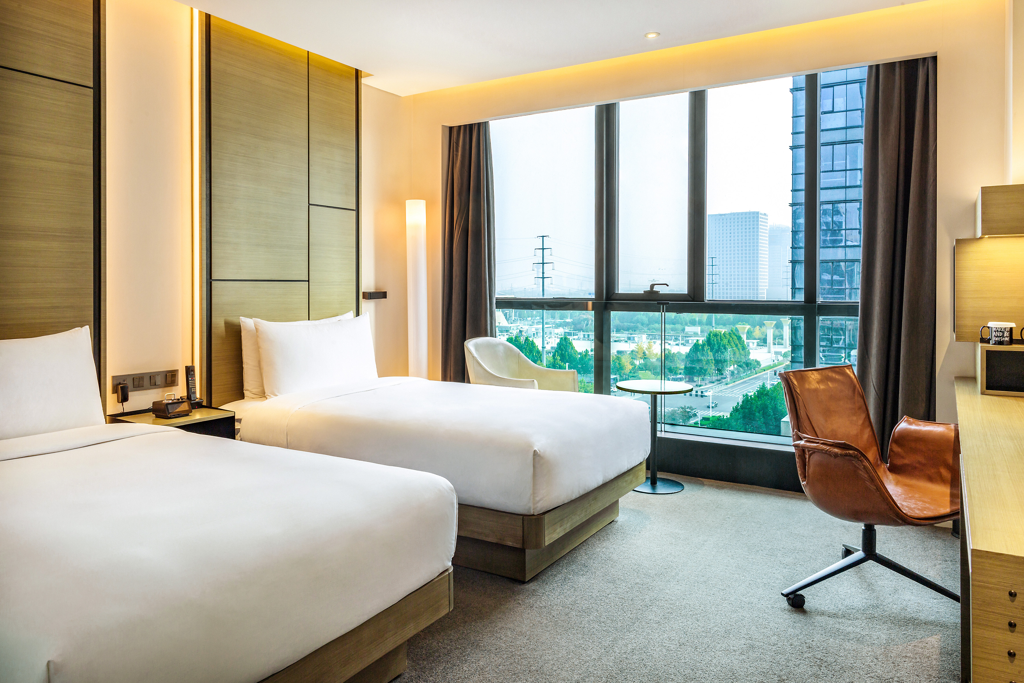 Contemporary hotel room with twin beds, cityscape view, and desk.