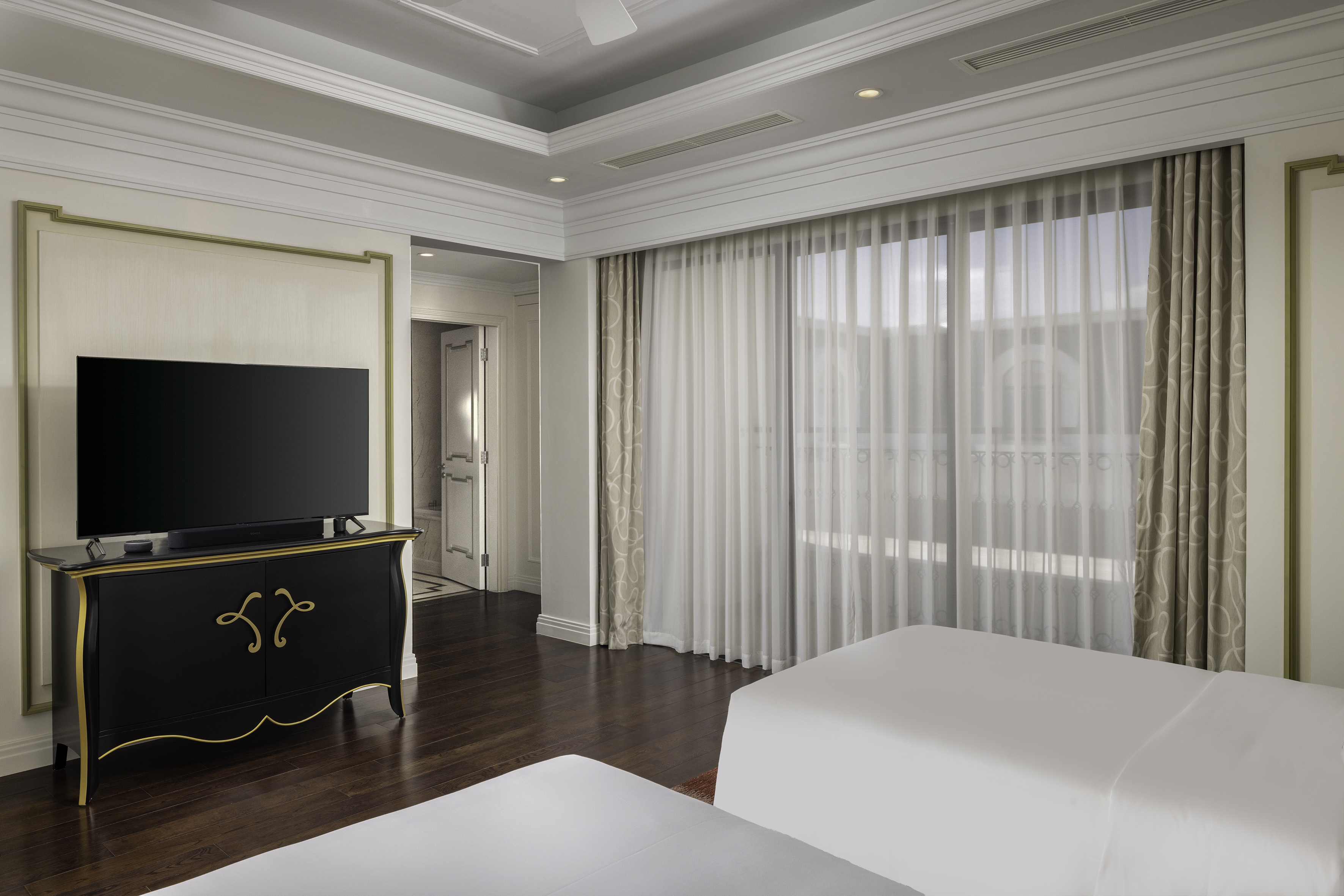 Luxury hotel bedroom with TV, dark wood floor, and large window with sheer curtains.