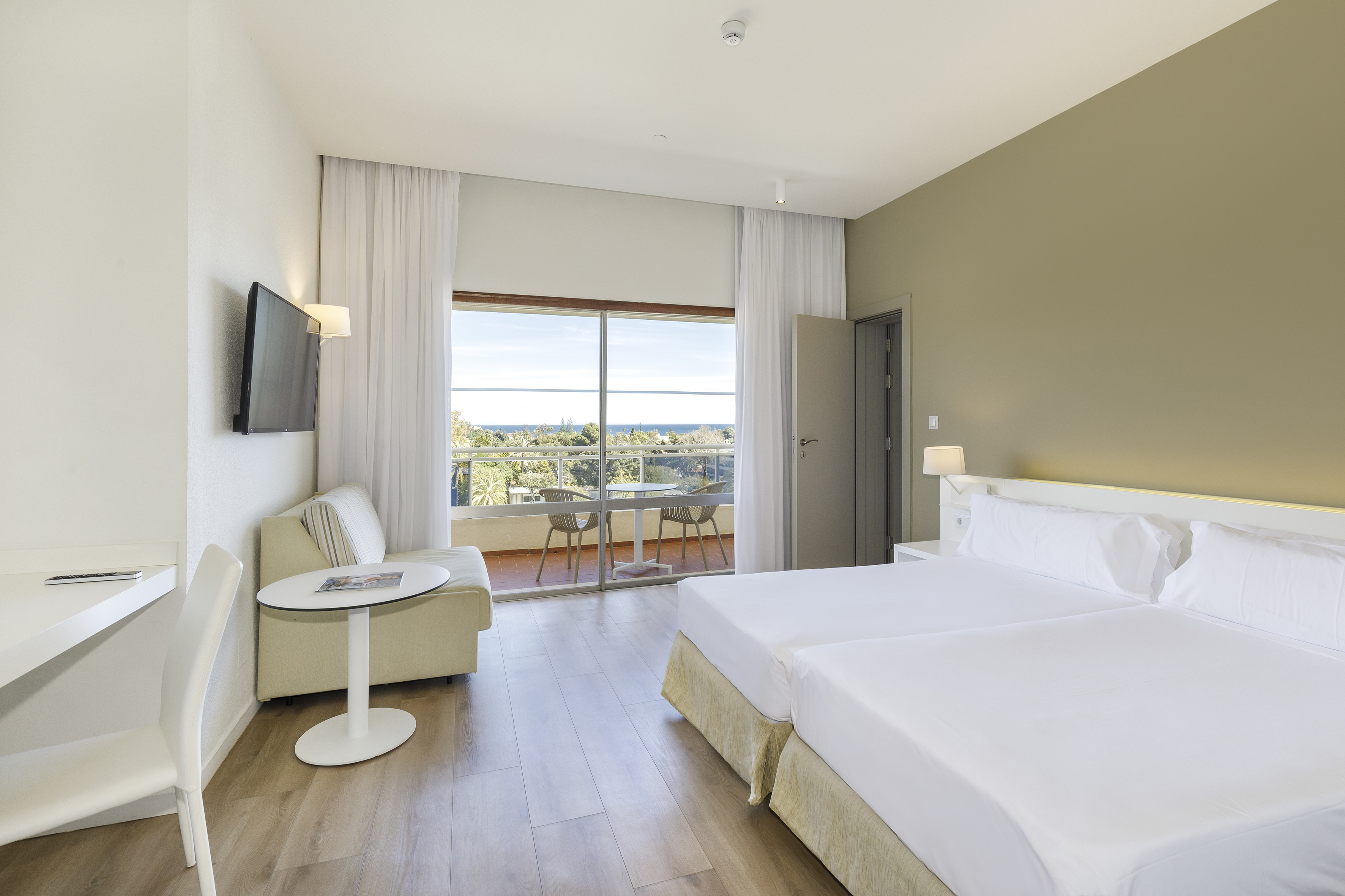 Modern hotel room with twin beds, balcony, ocean view, desk, and sofa.