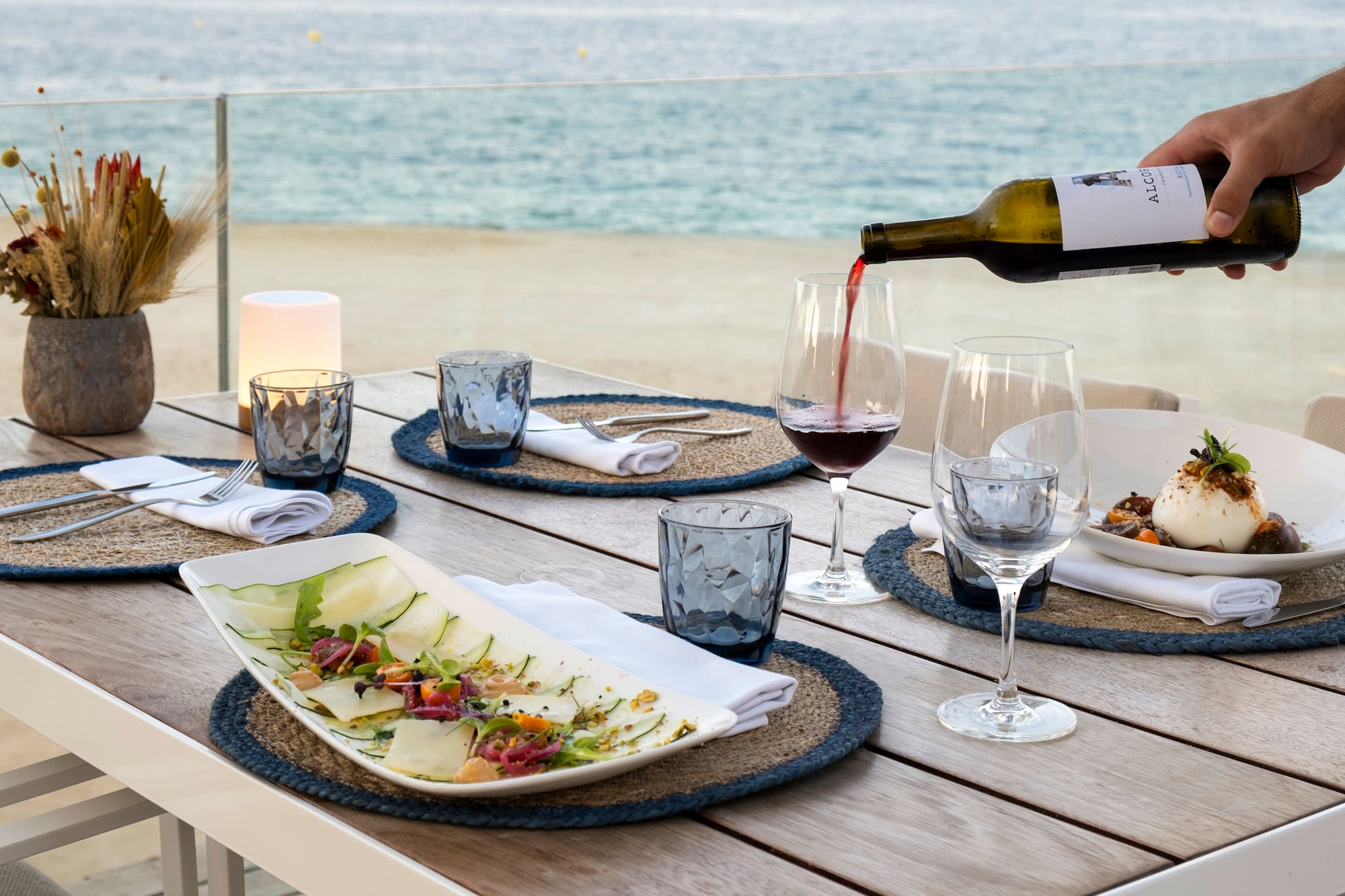 Mesa junto al mar con vino tinto, burrata y ensalada fresca.
