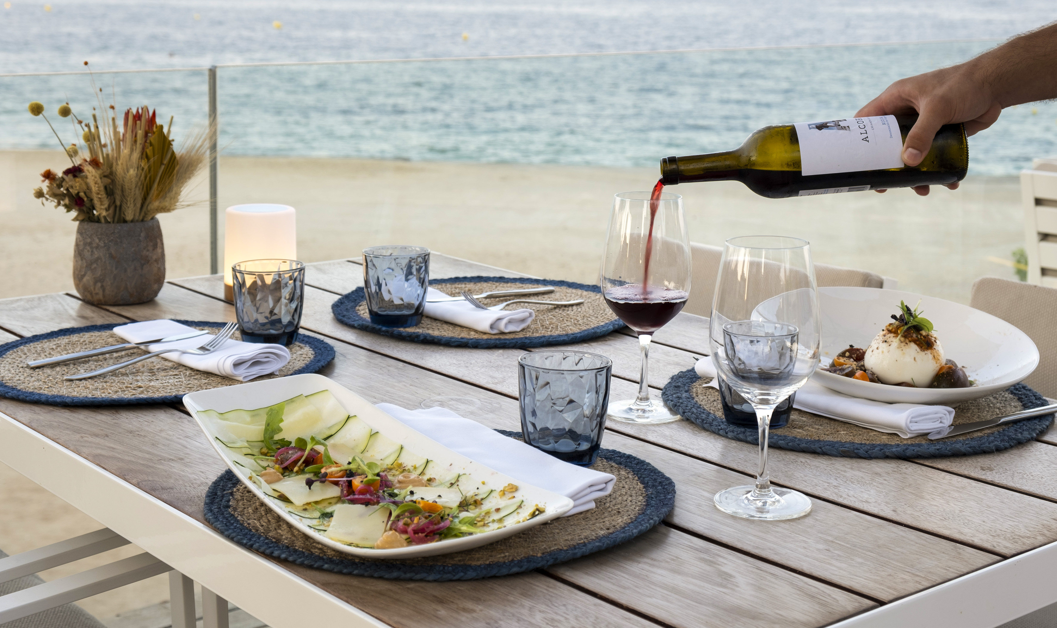 Mesa junto al mar con vino tinto, burrata y ensalada fresca.