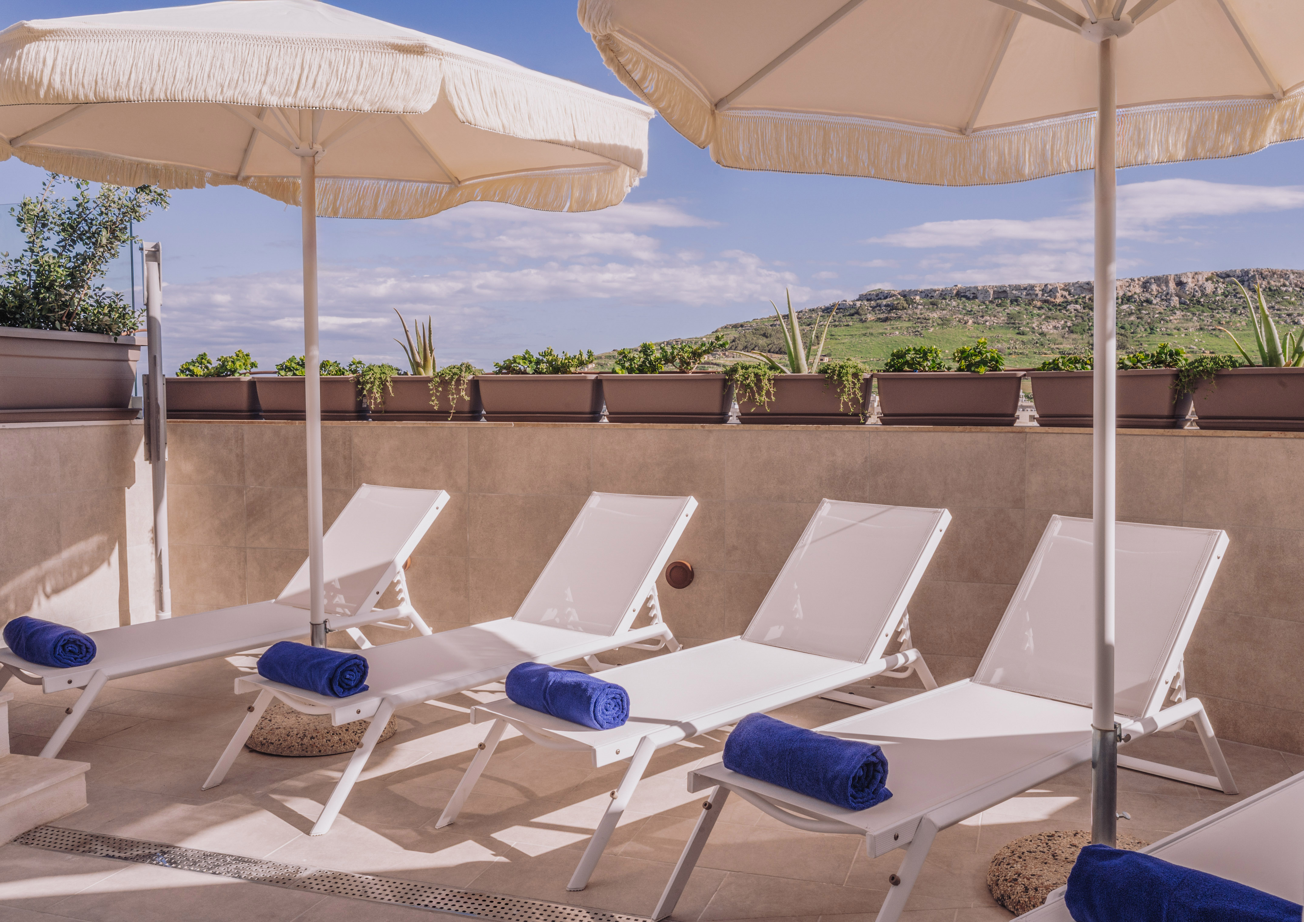 Outdoor patio with white sun loungers, blue towels, parasols, and green hill view.