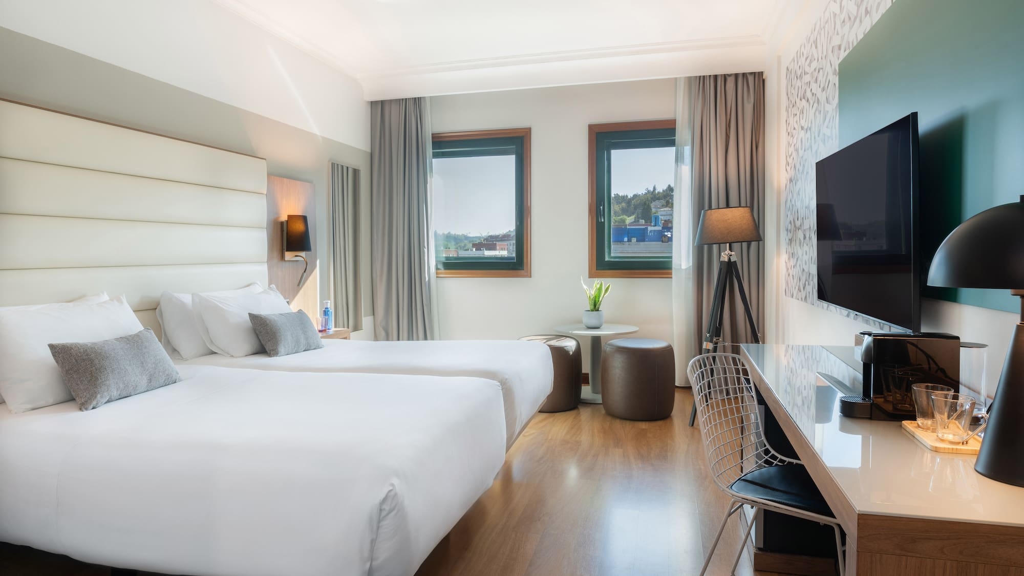 Modern hotel room with twin beds, large windows, desk, and TV.