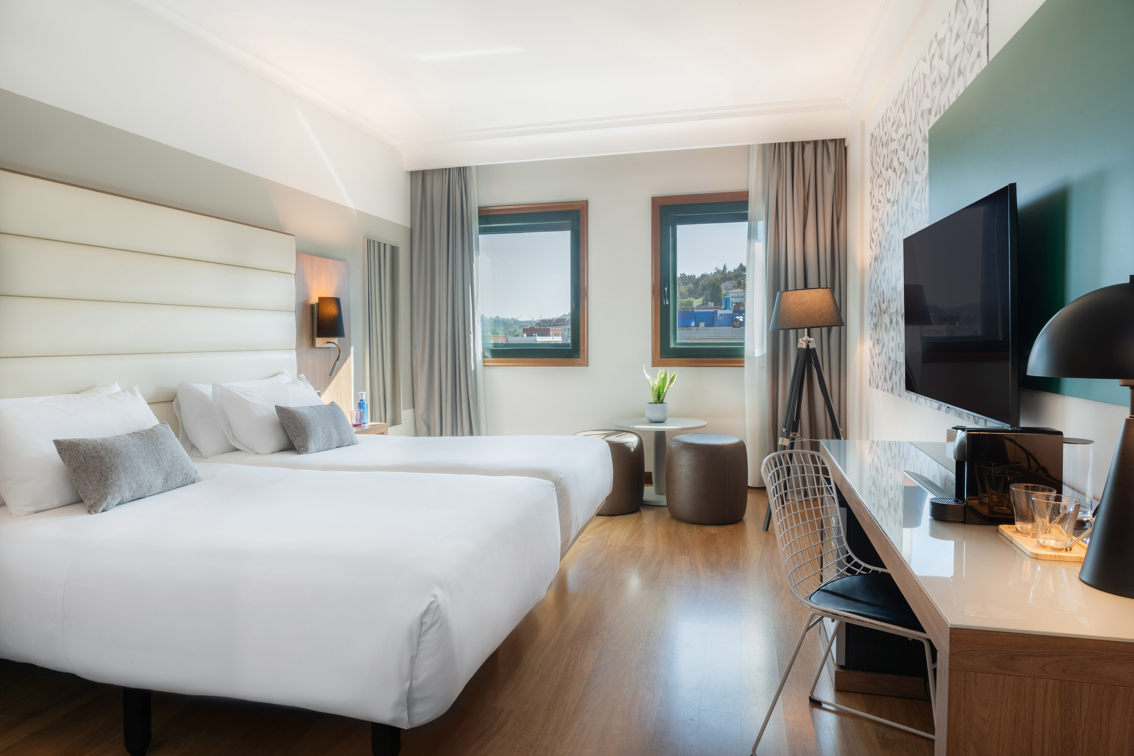 Modern hotel room with twin beds, large windows, desk, and TV.