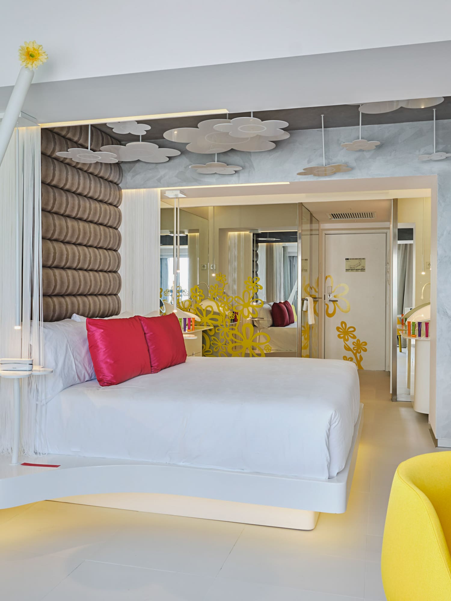 Modern hotel room: white bed, red pillows, yellow chair, unique decor.