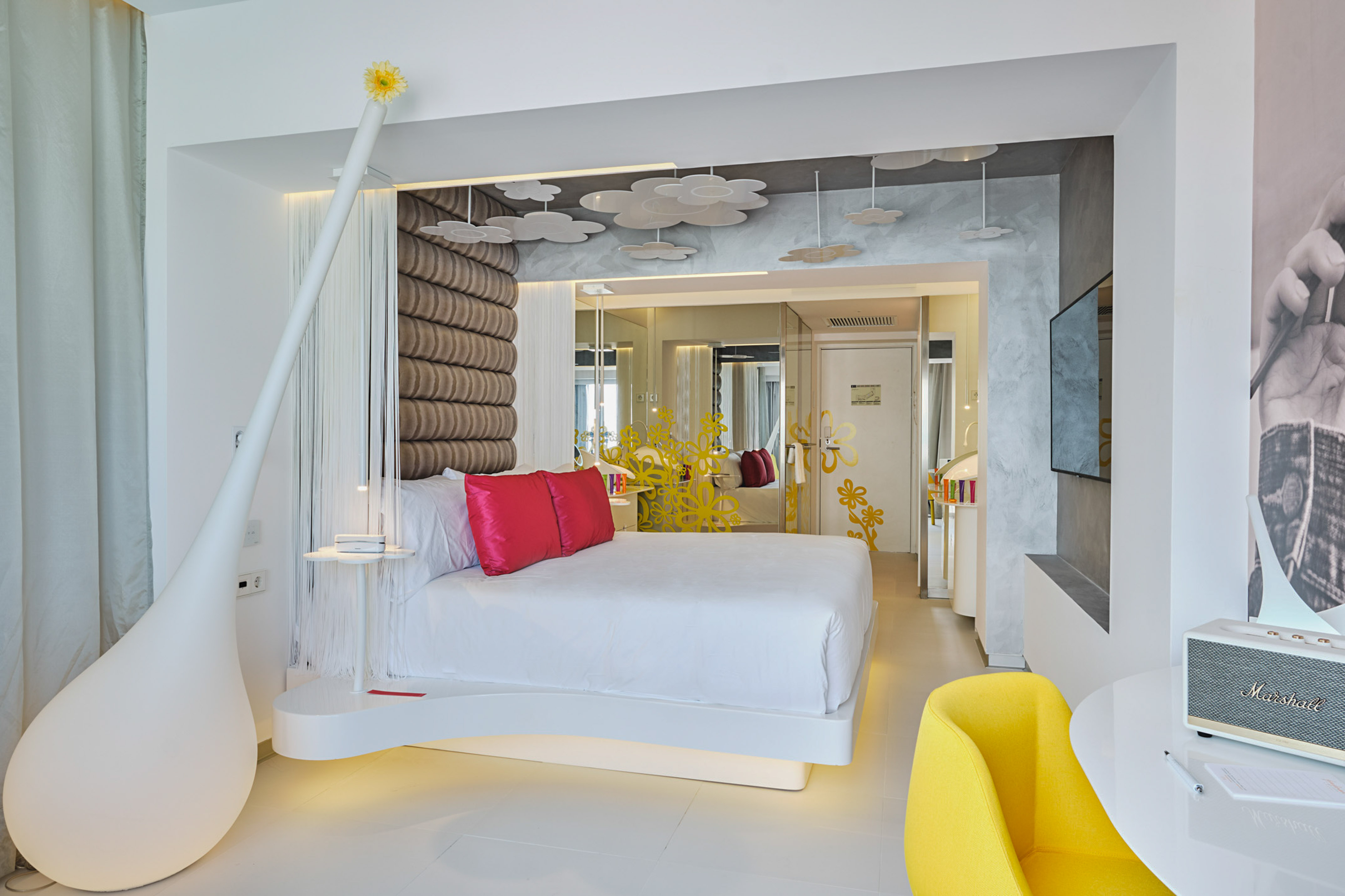 Modern hotel room: white bed, red pillows, yellow chair, unique decor.